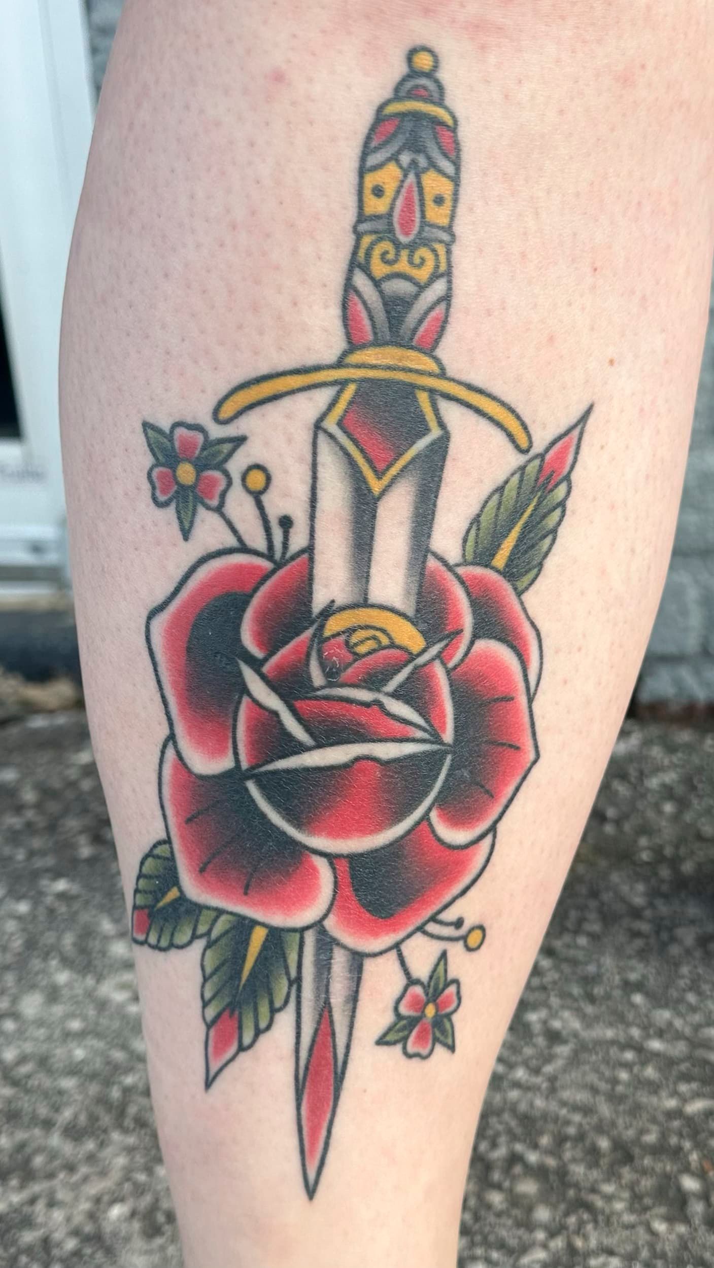 Tattoo of a dagger piercing a red rose with green leaves and small flowers