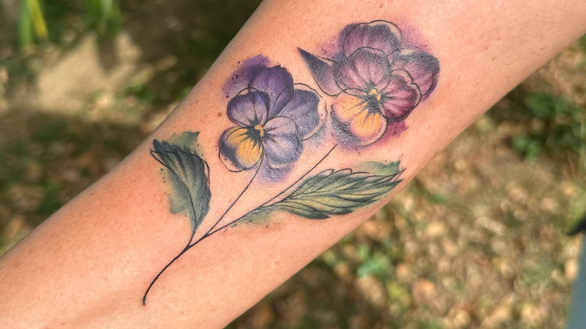 Purple flower tattoo with green leaves on a forearm