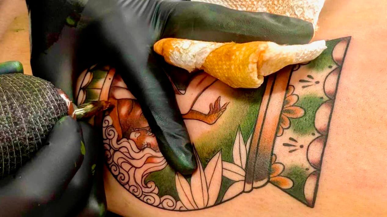 Gloved hands apply ointment to a colorful tattoo on skin with a tissue above it.