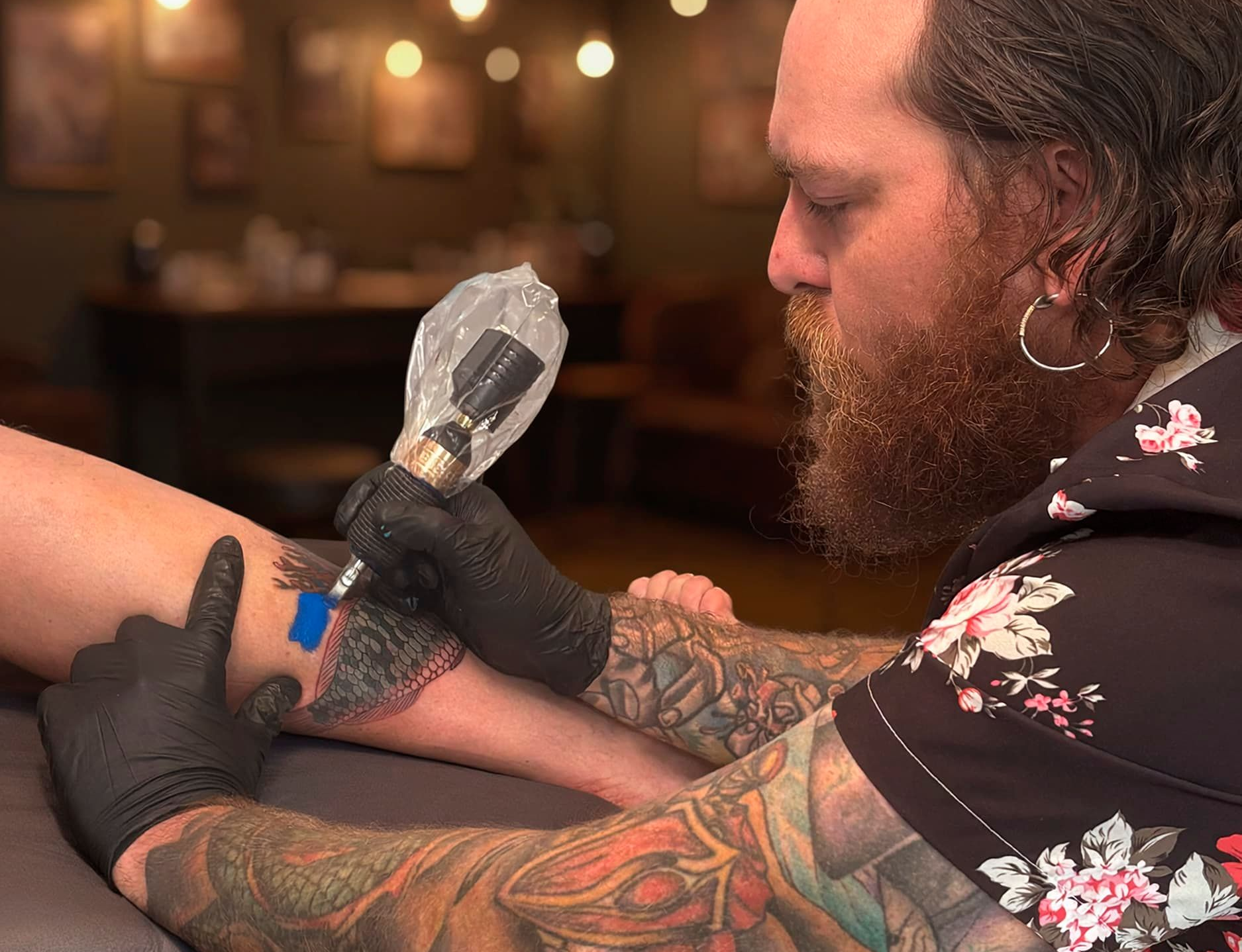 Tattoo artist in black gloves uses a tattoo machine on a client’s arm in a warm studio.