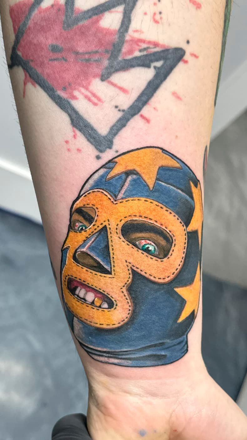 Close-up of a forearm tattoo of a colorful masked face with orange and blue details.