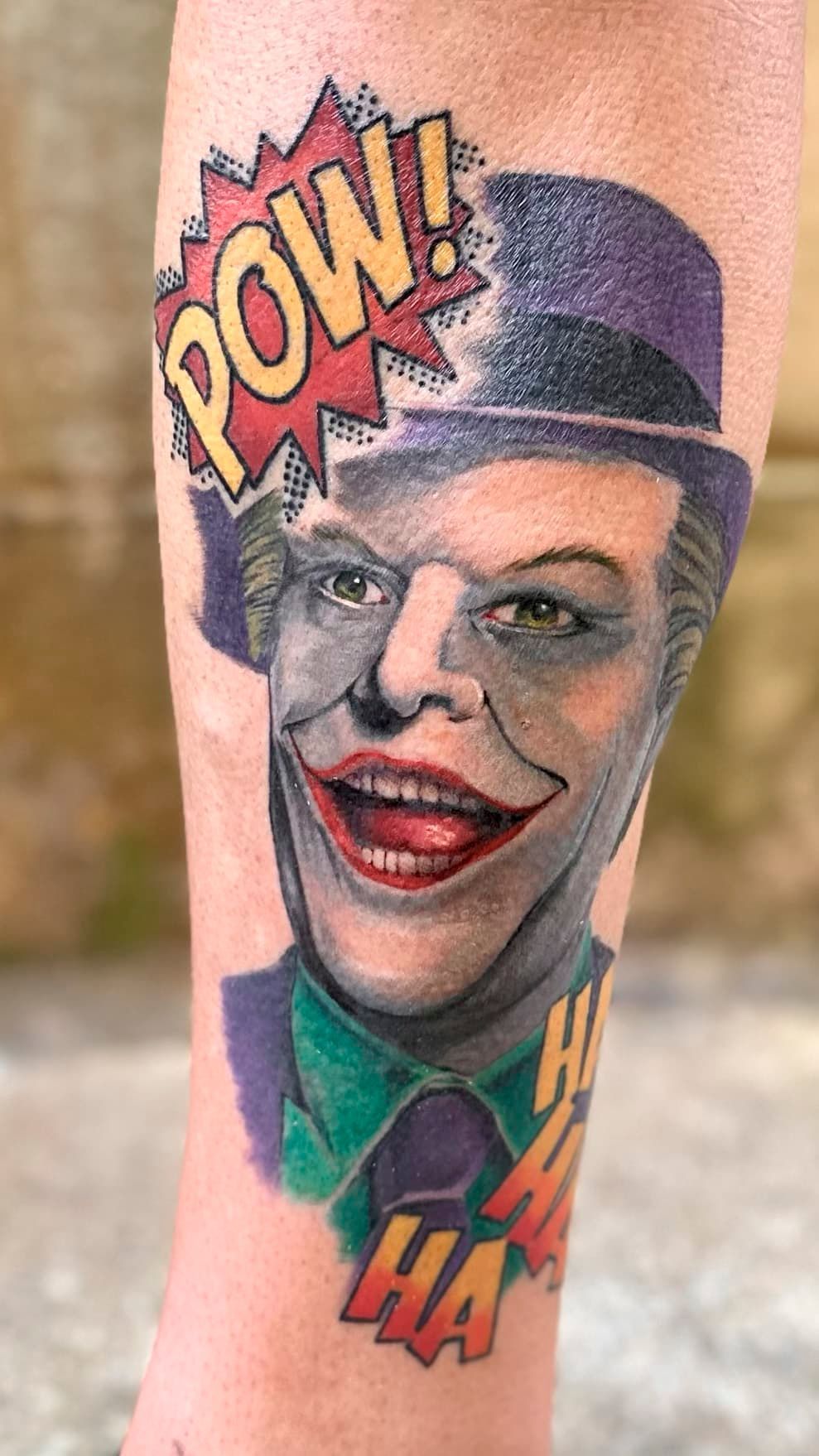 Colorful tattoo of a grinning comic-style face with a hat, speech bubbles saying “POW!” and “HA”