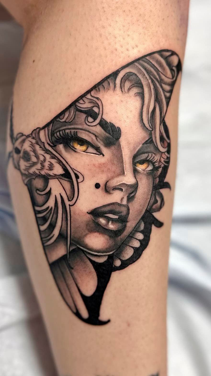 Black-and-gray tattoo of a woman’s face with ornate swirling details on a leg