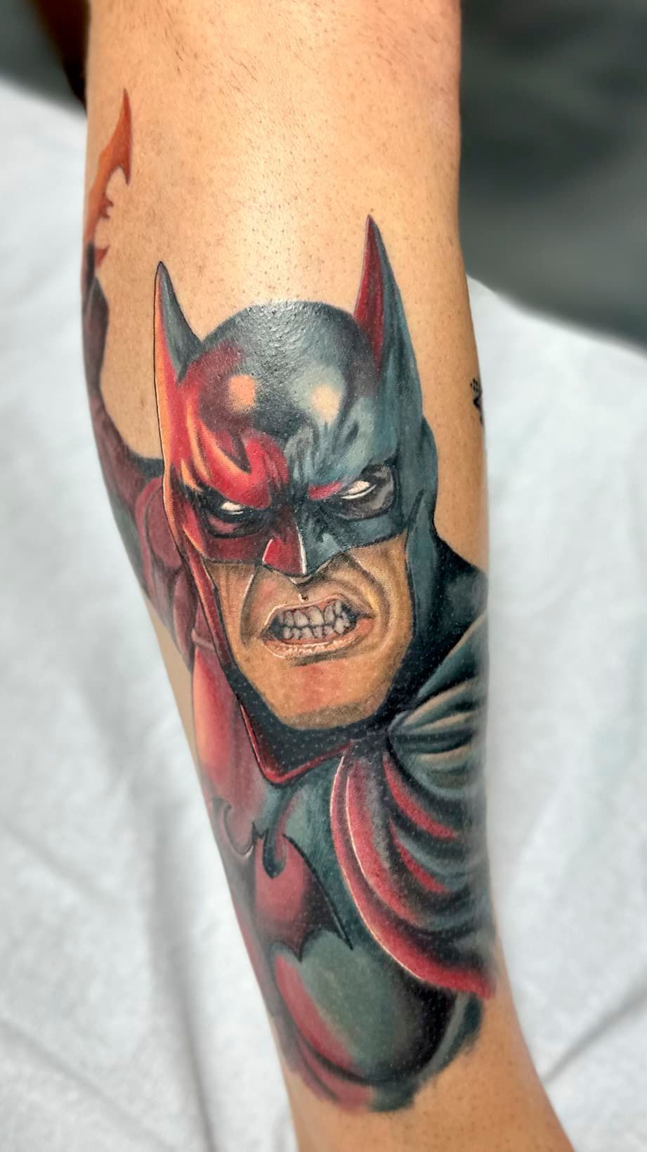 Colorful Batman tattoo on a forearm, showing a fierce masked face and red-black costume details.