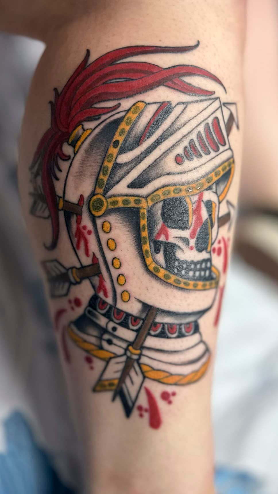 Colorful tattoo of a skull with a red mohawk and feather, wrapped in gold beads on a forearm