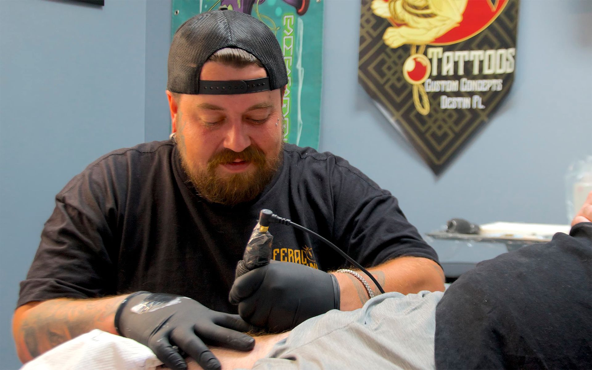 Tattoo artist in black gloves working at a studio table, with a tattoo shop sign in the background