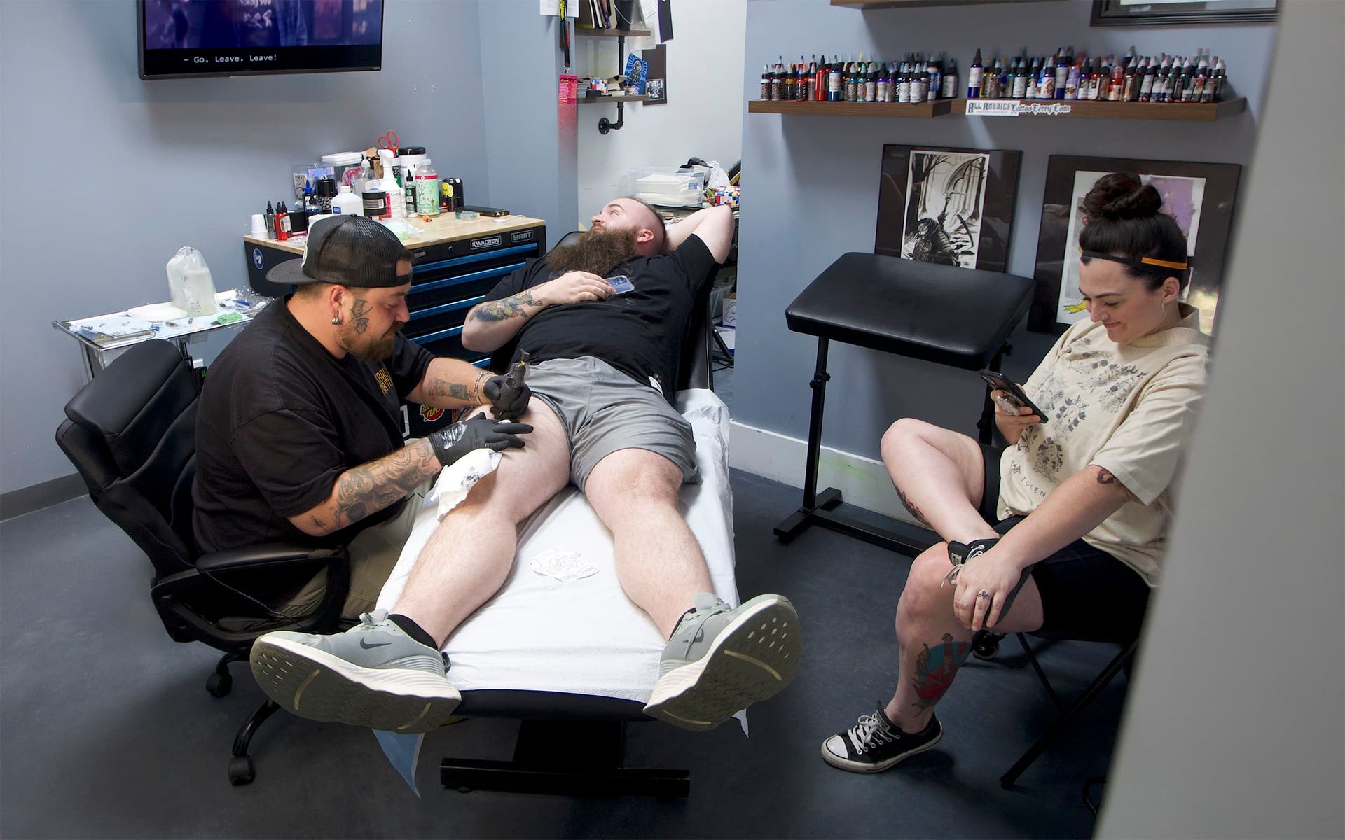 Tattoo artist working on a client’s leg while another person watches in a tattoo studio
