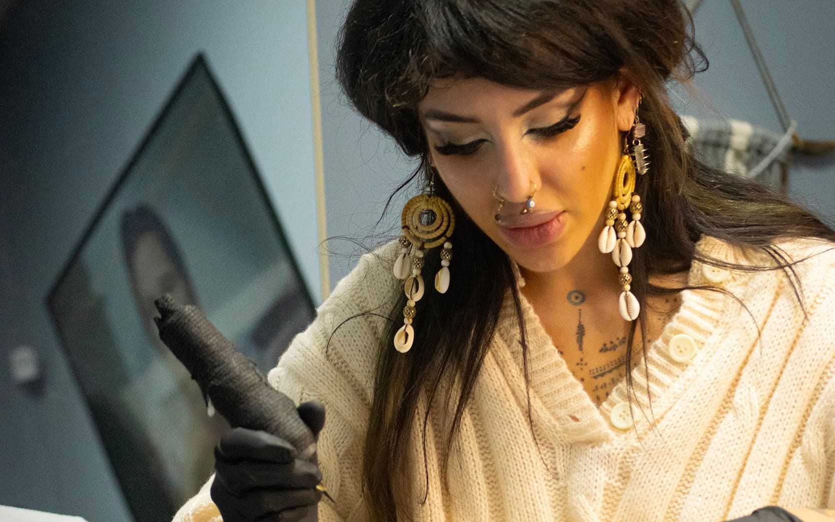 Woman in cream sweater and black gloves examining an object indoors, with a framed artwork in the background