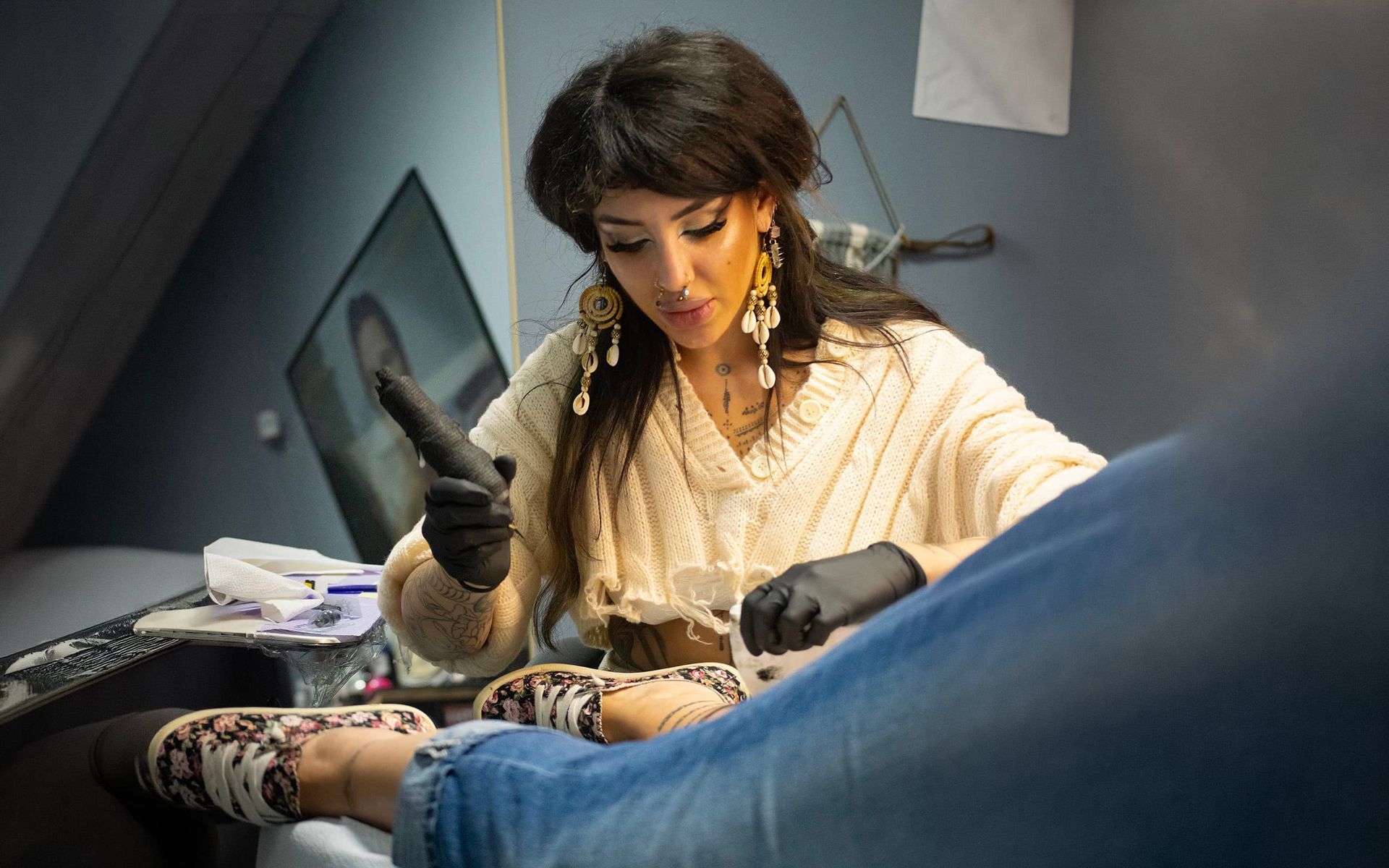 Tattoo artist in black gloves working on a client’s leg in a studio, with sketches on the wall.
