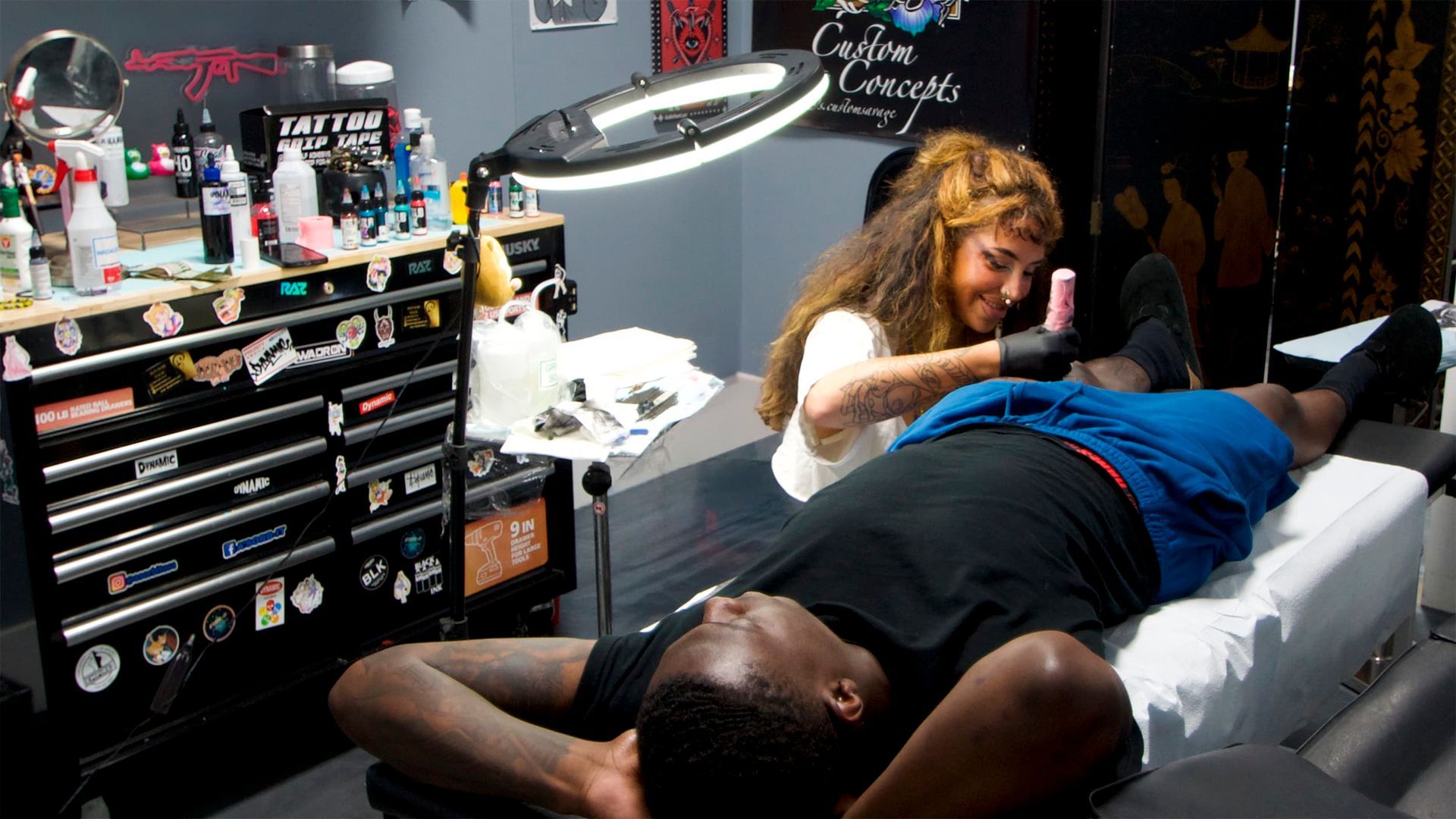Tattoo artist working on a client’s back in a studio, with supplies and a task lamp nearby