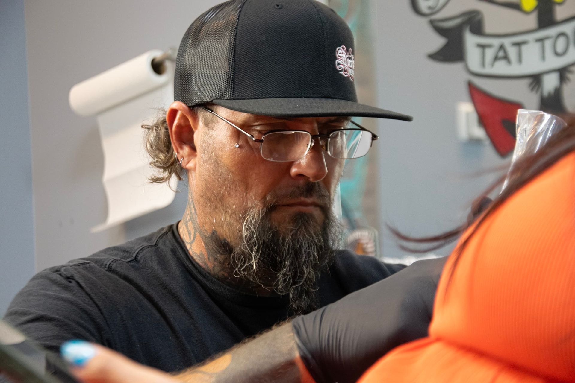 Tattoo artist in black cap working on a client’s arm, focused in a studio with a tattoo sign in the background