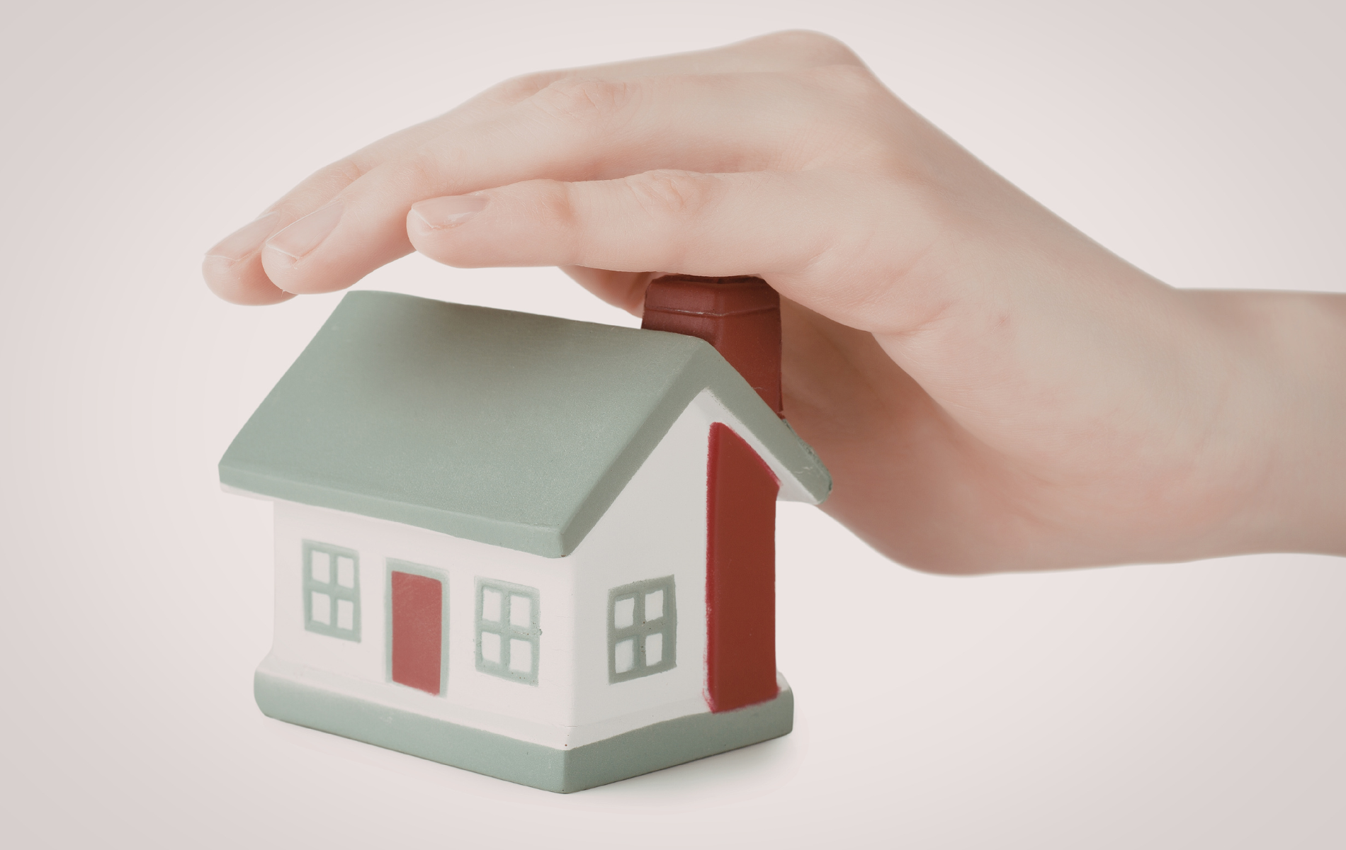 A hand held protectively over a small model house with a green roof and a red chimney.