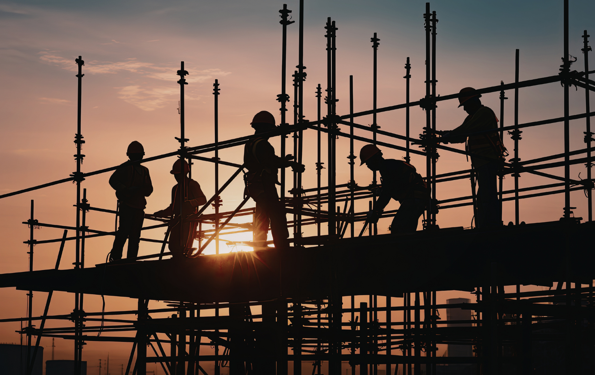 Silhouetted construction workers on scaffolding against a bright sunset sky.