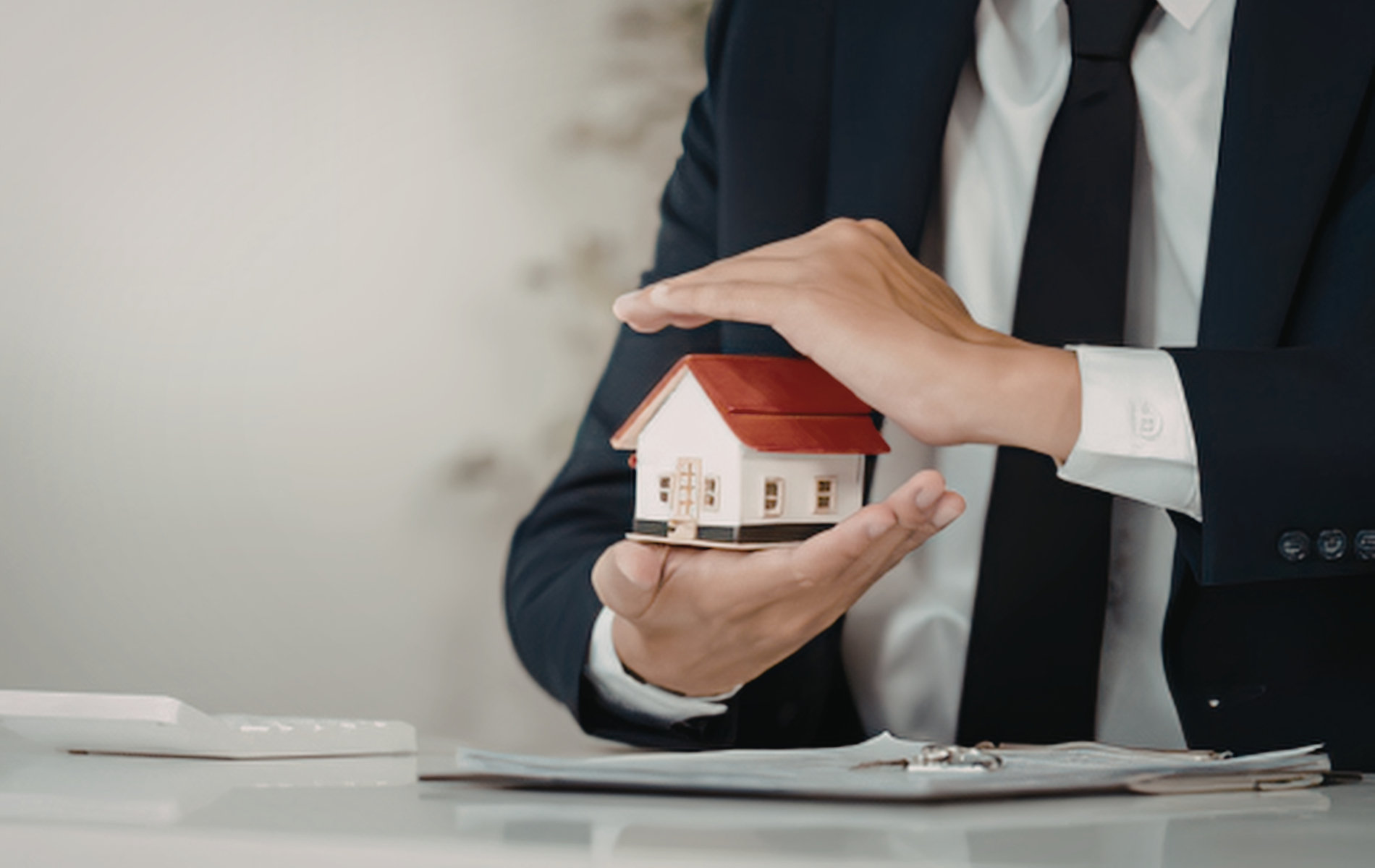 A person in a business suit protects a miniature house model with their hands.