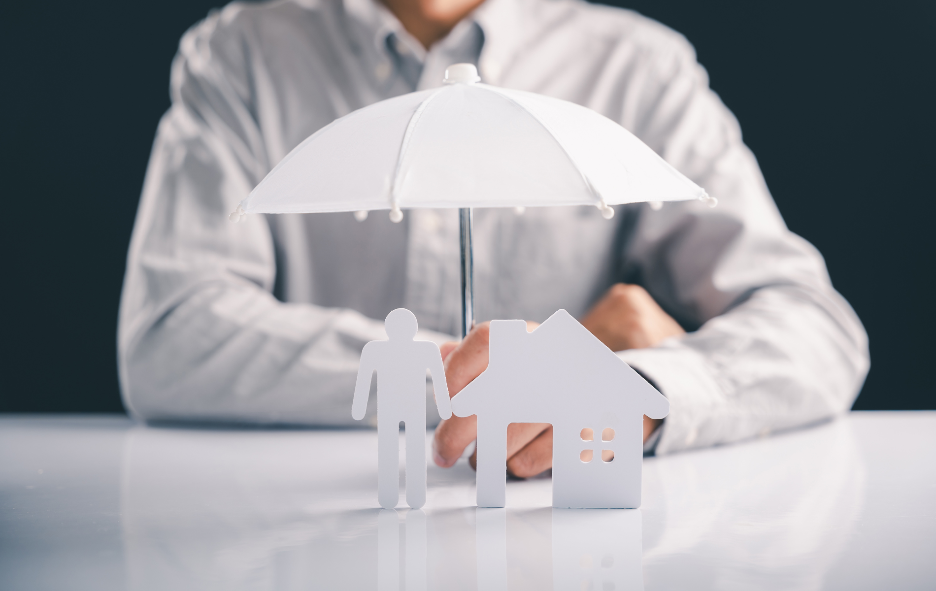 A person in a light shirt sits behind a desk holding an umbrella over a white paper cutout of a house and a human figure.
