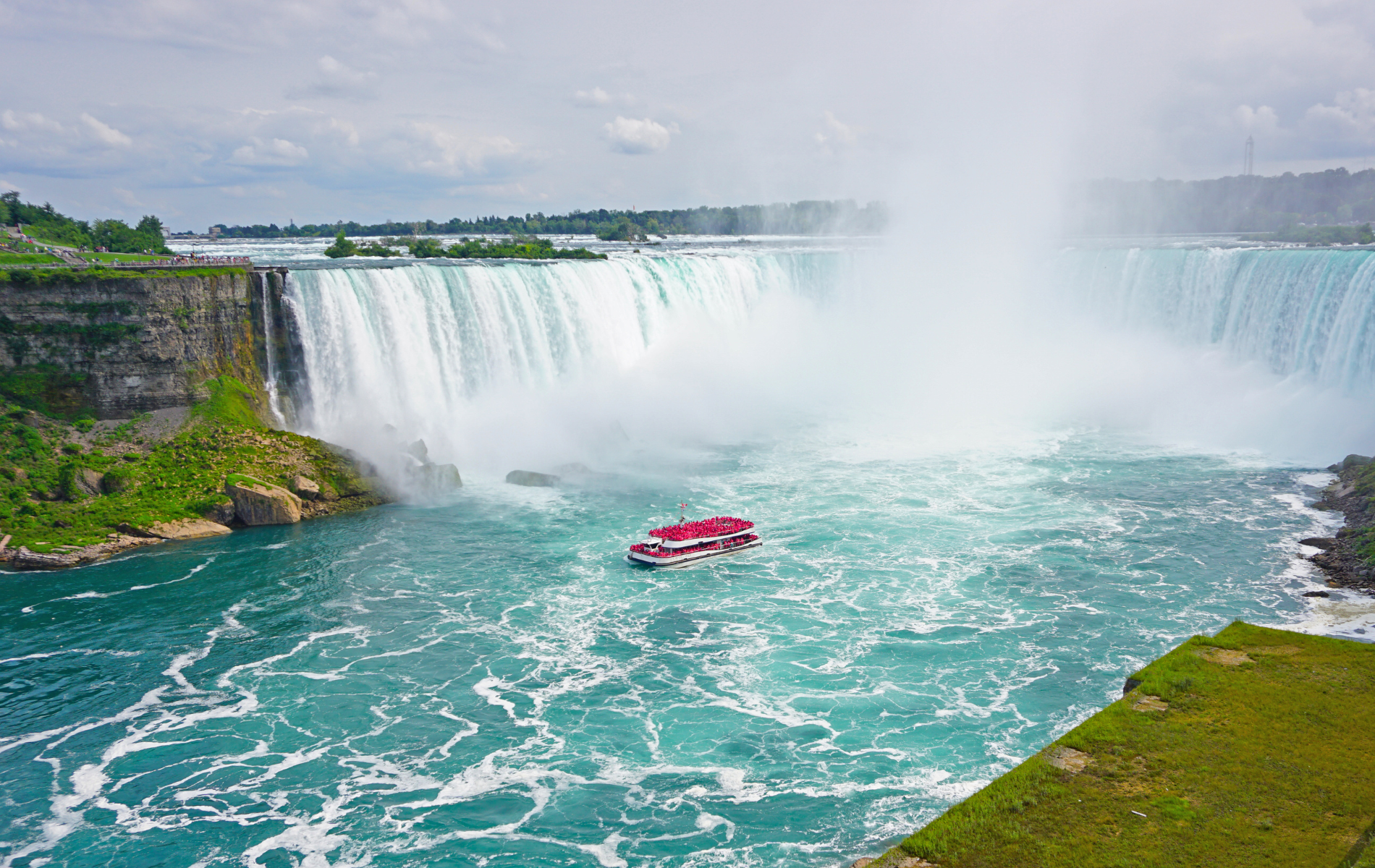 A red tour boat navigates the turbulent turquoise water at the base of the massive Horseshoe Falls at Niagara Falls.