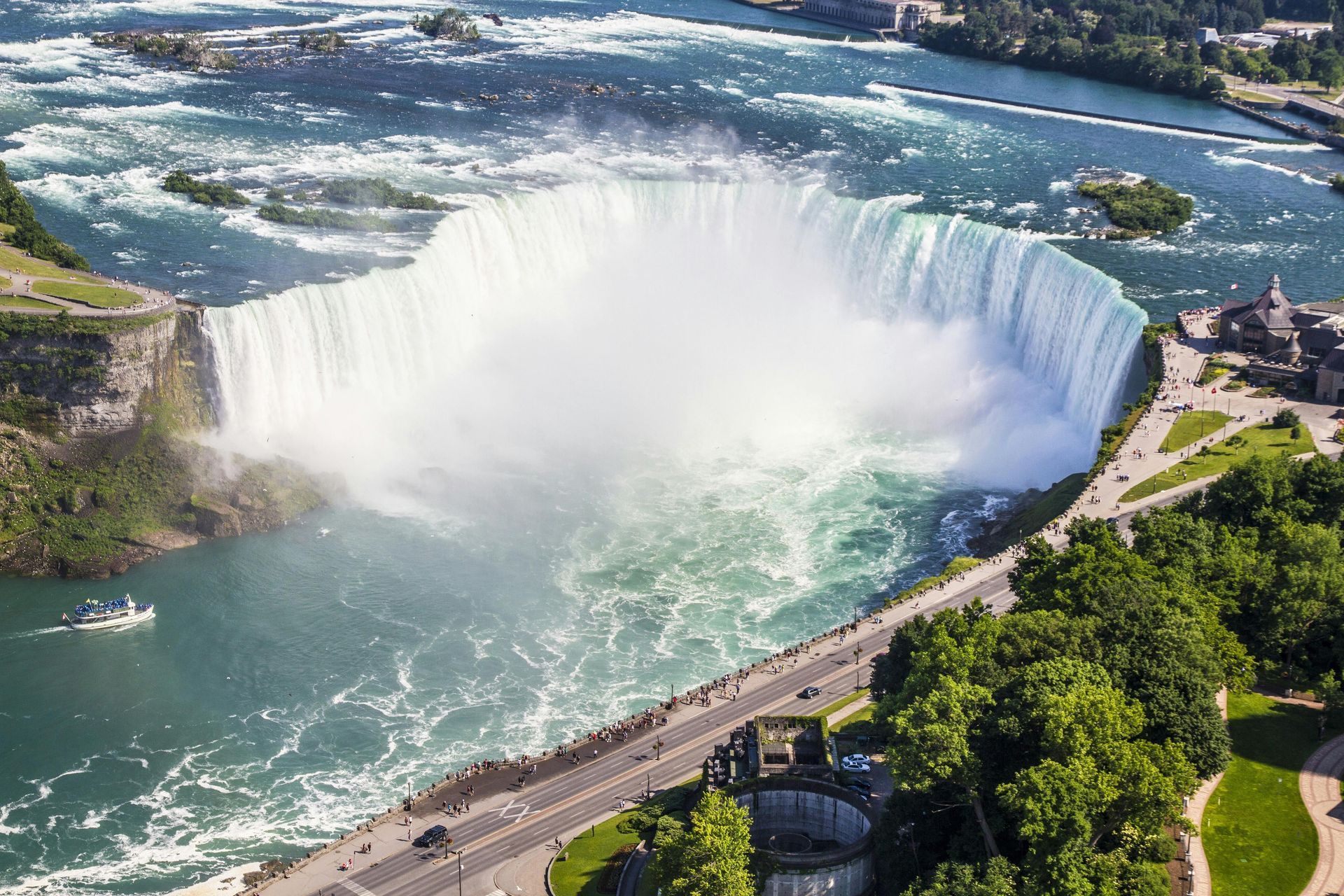Aerial view of the Horseshoe Falls at Niagara Falls, featuring a mist-filled basin with a road and parkland alongside.