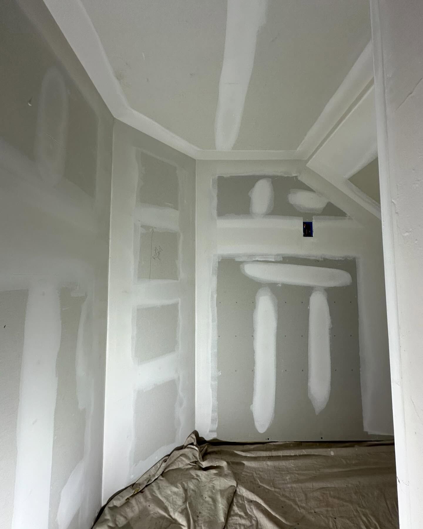 Interior room under construction, with drywall and taped seams. Brown tarp covers floor. White walls.