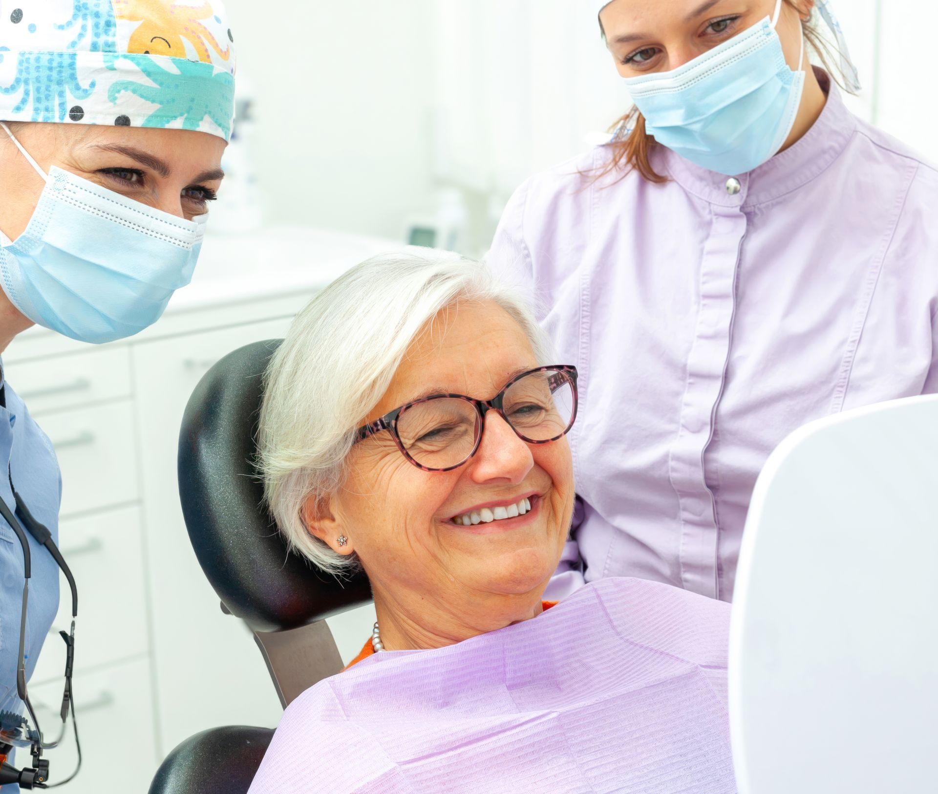 Smiling dental patient in chair with two masked clinicians in a bright clinic