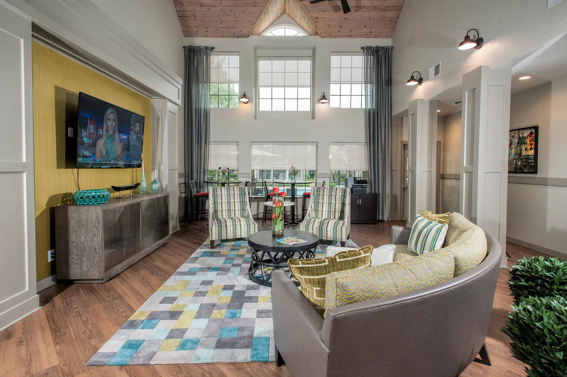 Bright community lounge with sofa, two striped chairs, a wall-mounted TV, and large windows at The Reserve at Capital Center.