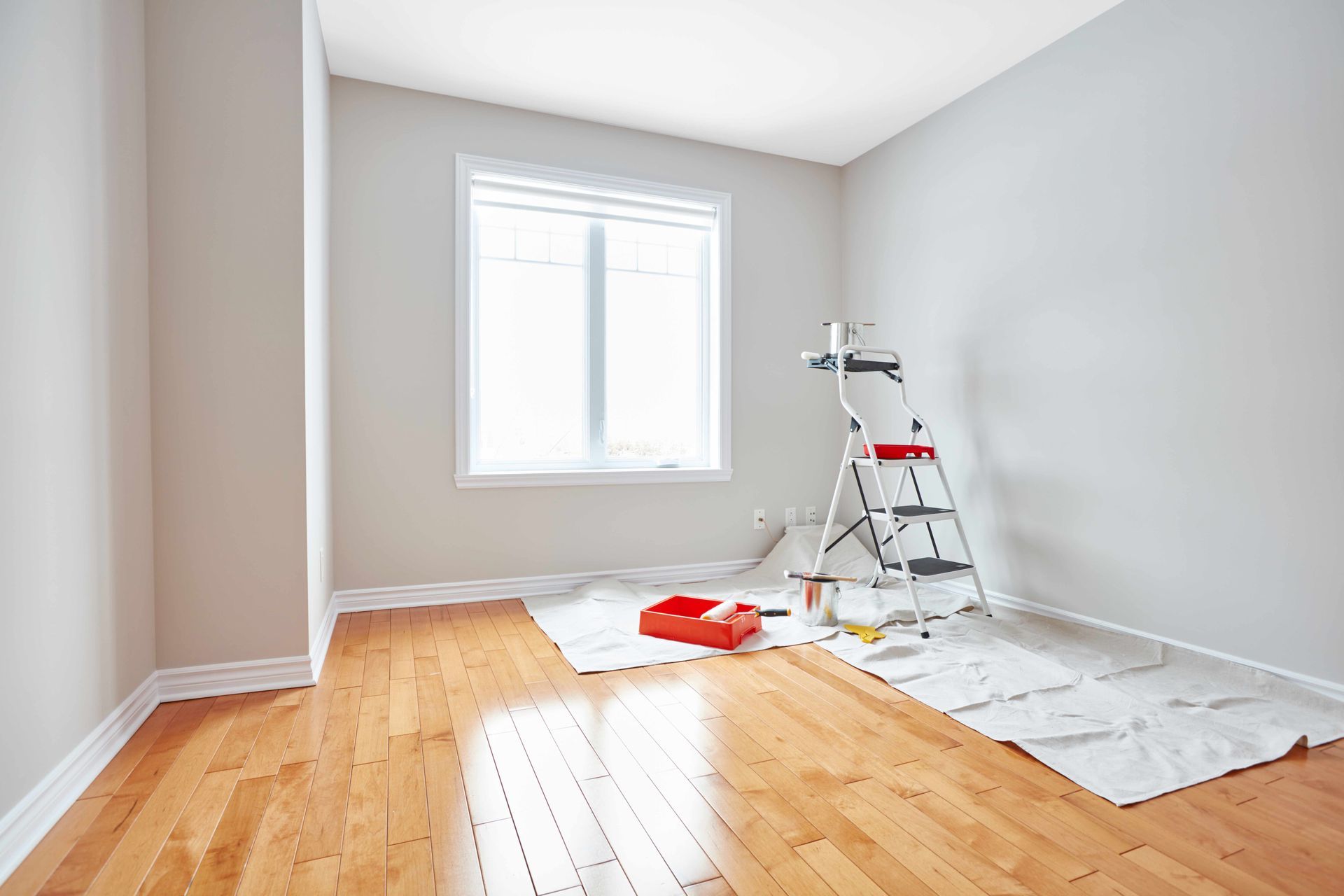 Empty room being painted, with a stepladder, drop cloths, and paint supplies on a hardwood floor. Empty room being painted, with a stepladder, drop cloths, and paint supplies on a hardwood floor.