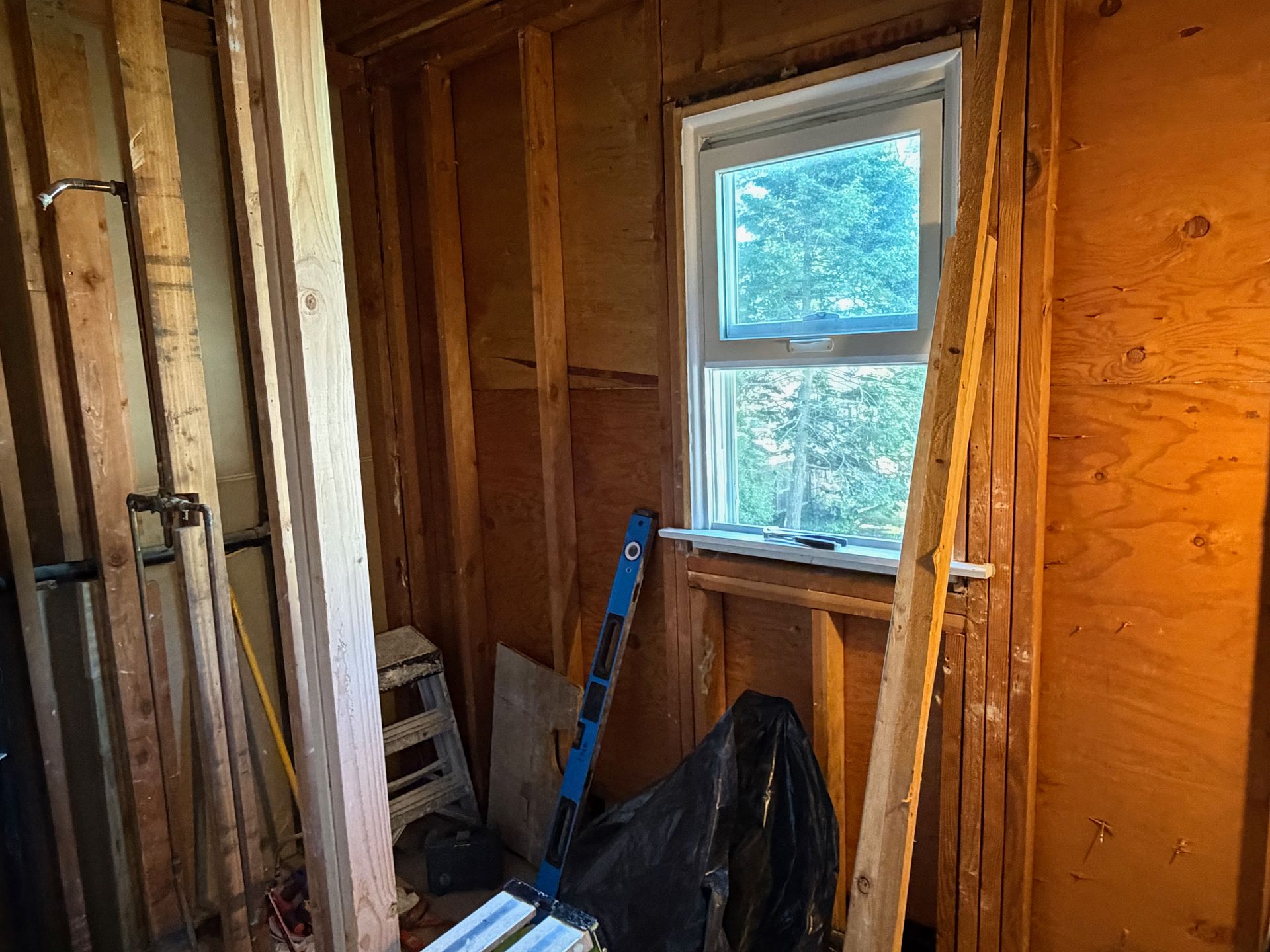 Interior of a bare wooden room under renovation, with exposed studs, a window, and tools leaning against the wall