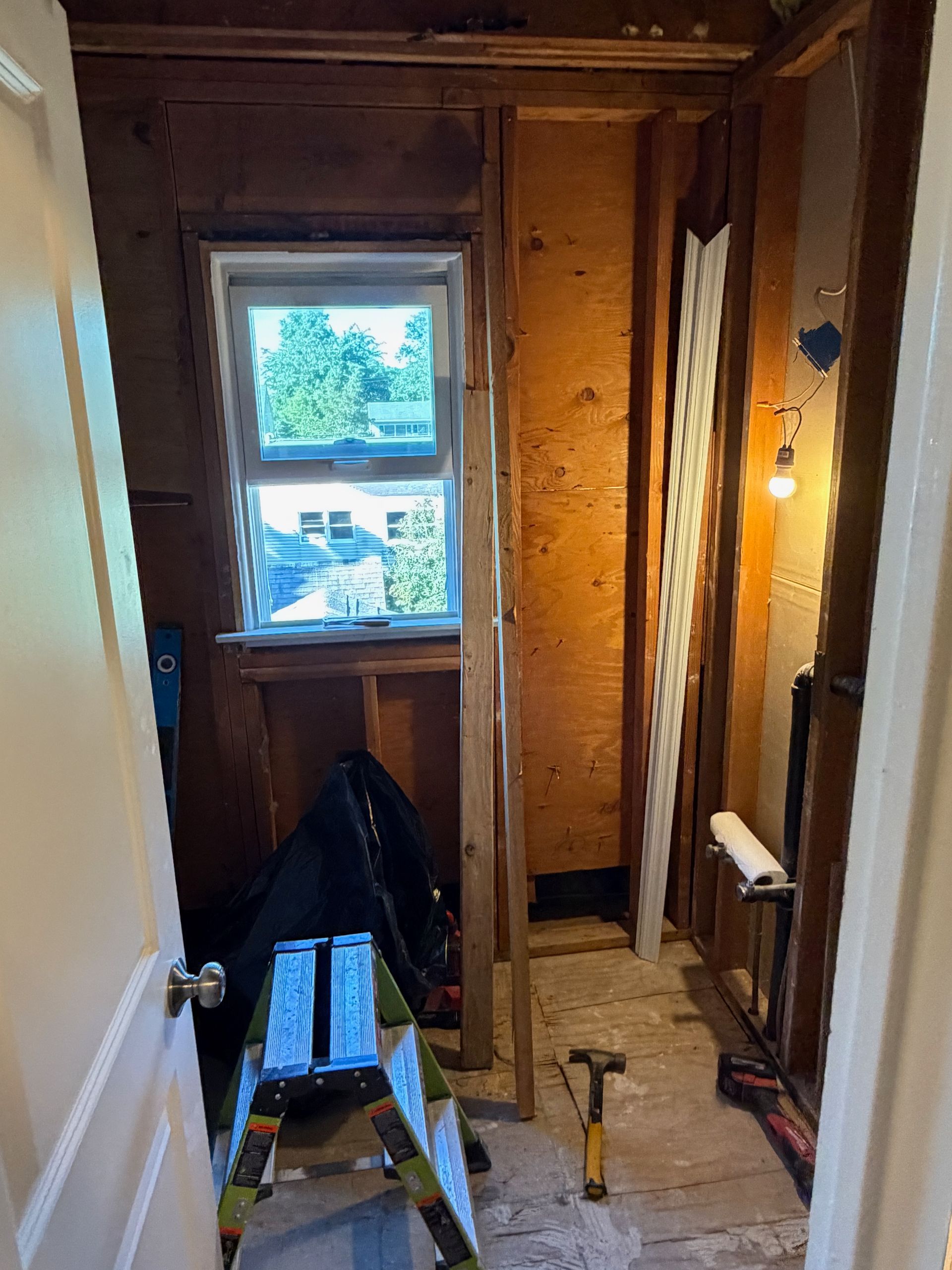 Narrow unfinished hallway under renovation with exposed wood, window, ladder, and tools on the floor