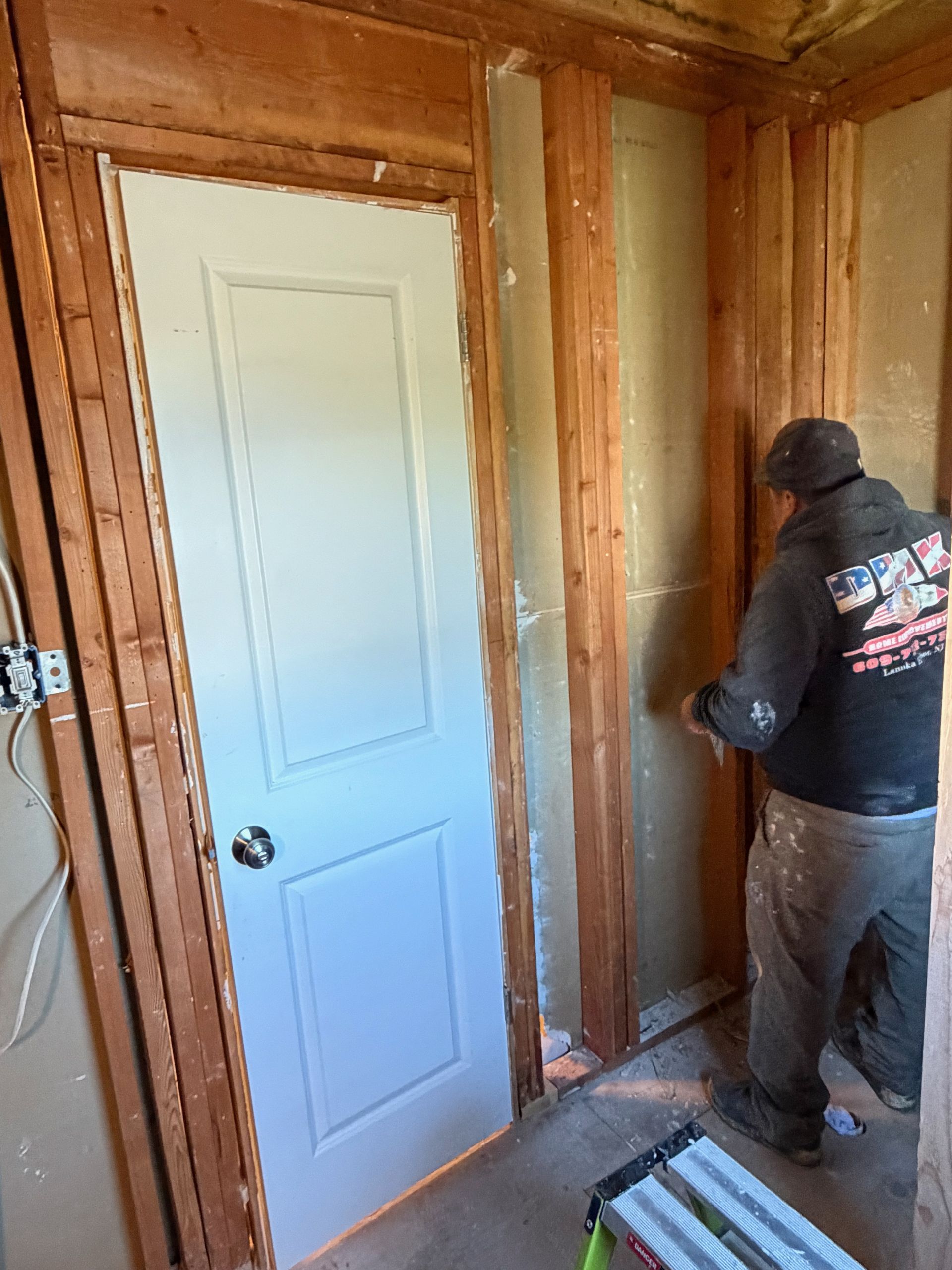 Worker installing drywall beside a white door in an unfinished room with exposed studs.