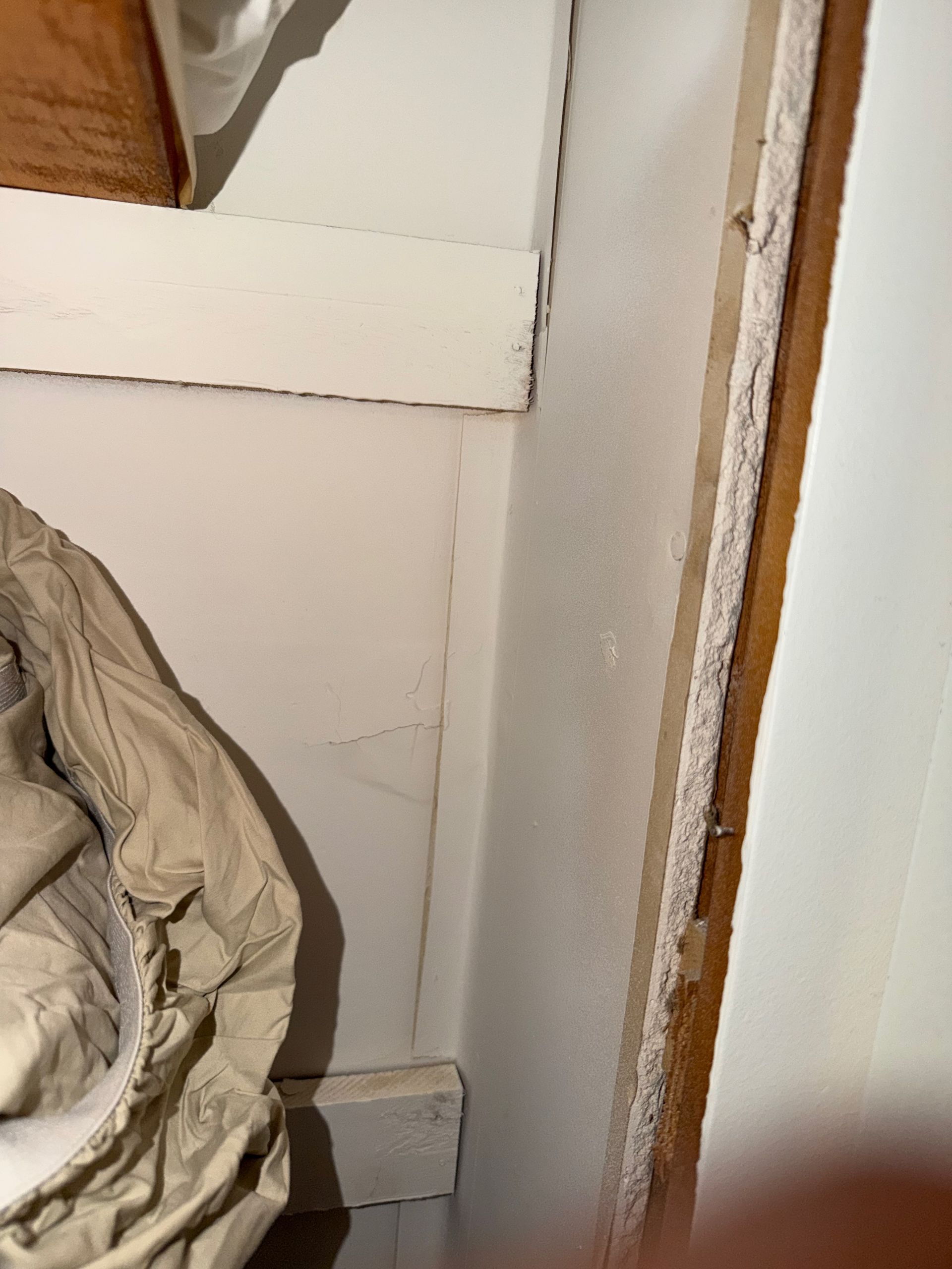 Corner of a small room with a beige crumpled blanket, white wall paneling, and wooden trim.