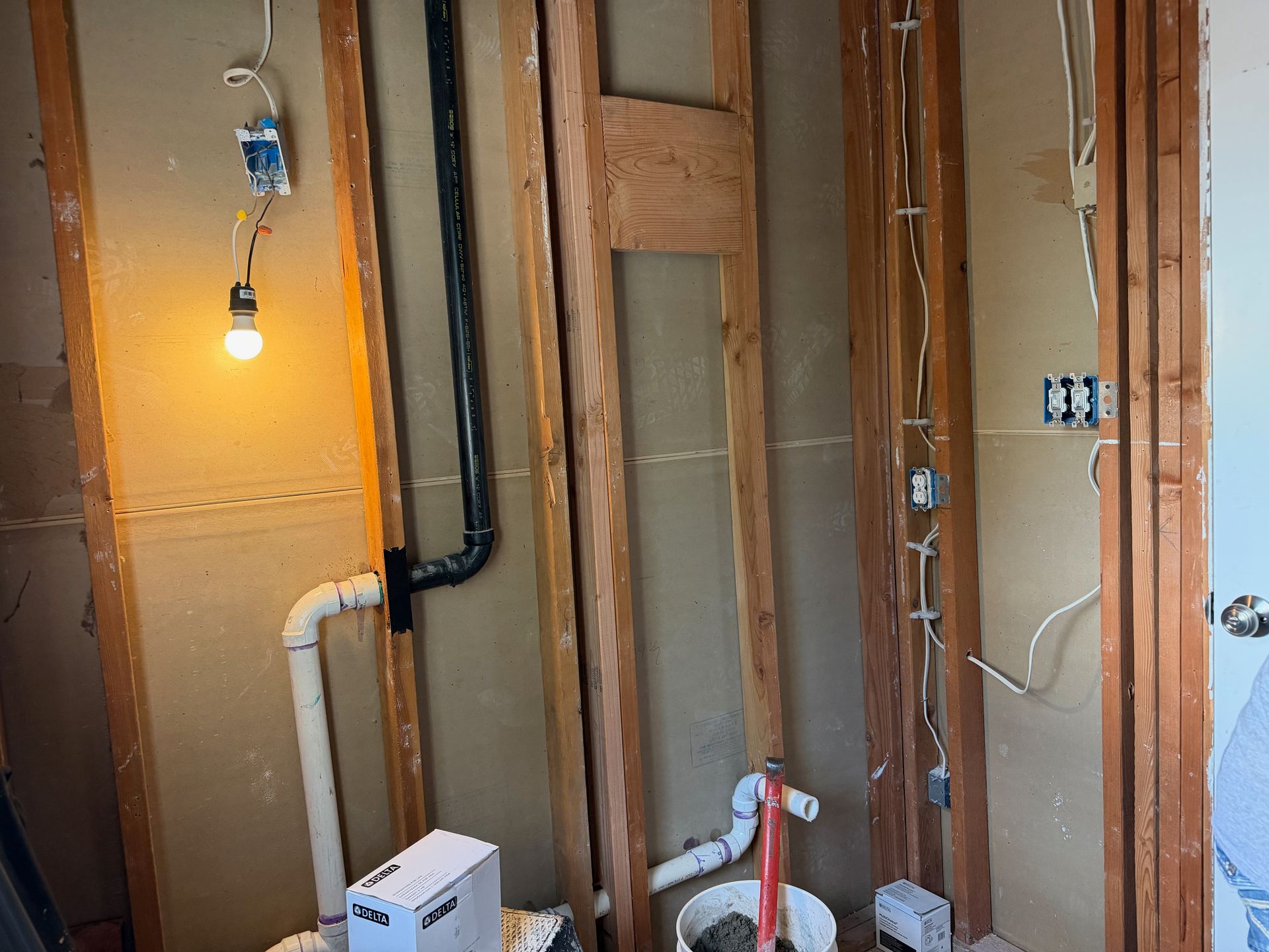 Exposed wall studs and plumbing in a bathroom under renovation with a hanging light bulb.