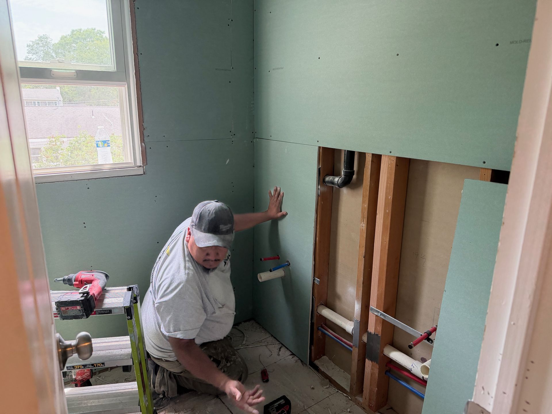 Worker installing plumbing in a small bathroom with open wall studs and green drywall.