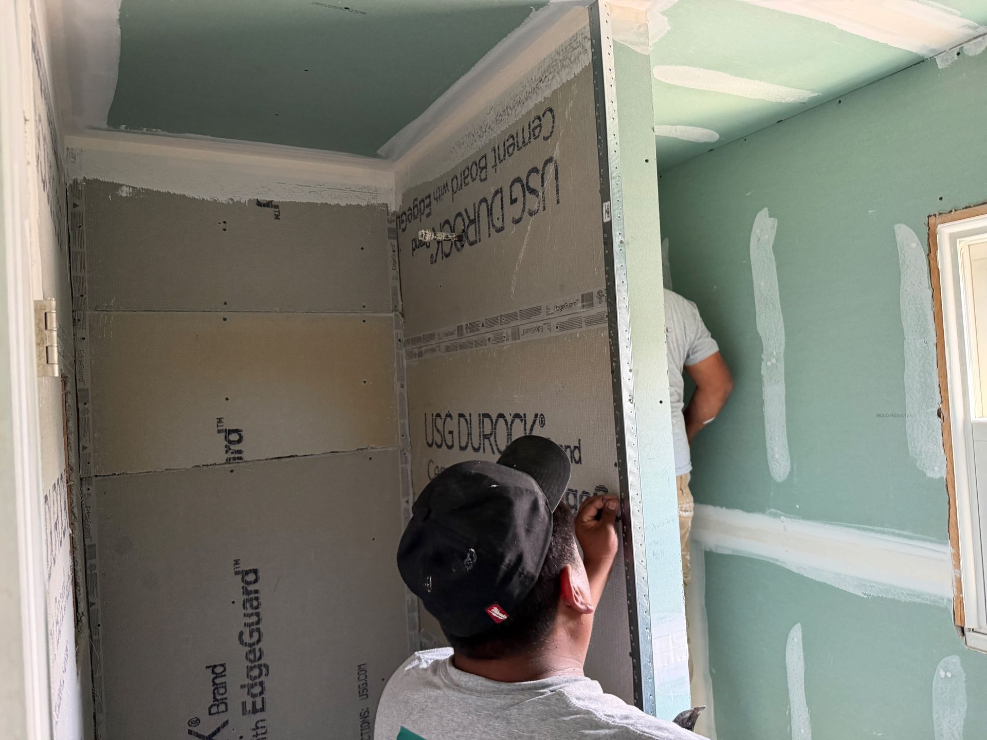 Two workers installing drywall in a room under renovation, with green walls and a window on the right.
