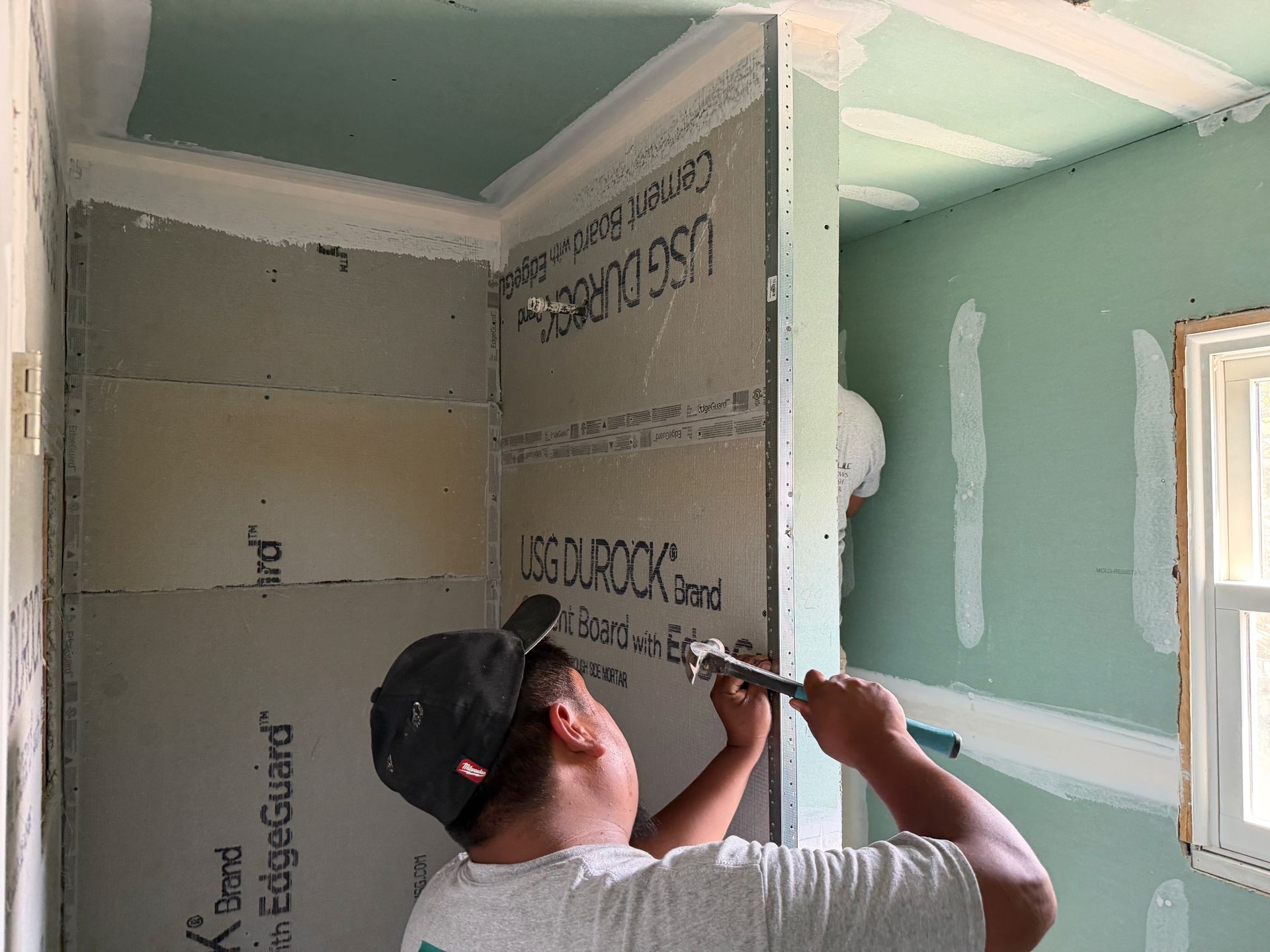 Worker installing drywall in a room, using a drill near a window and insulated wall panels.