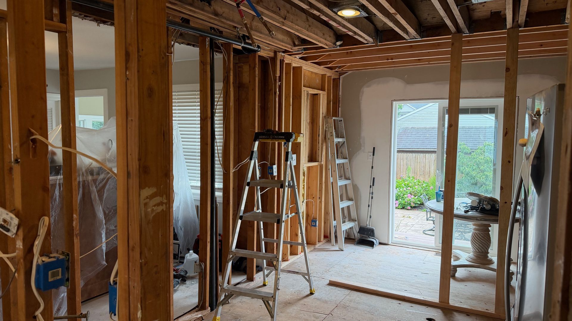 Renovation site with exposed wooden framing, ladder, and debris on the floor, looking into a bright doorway