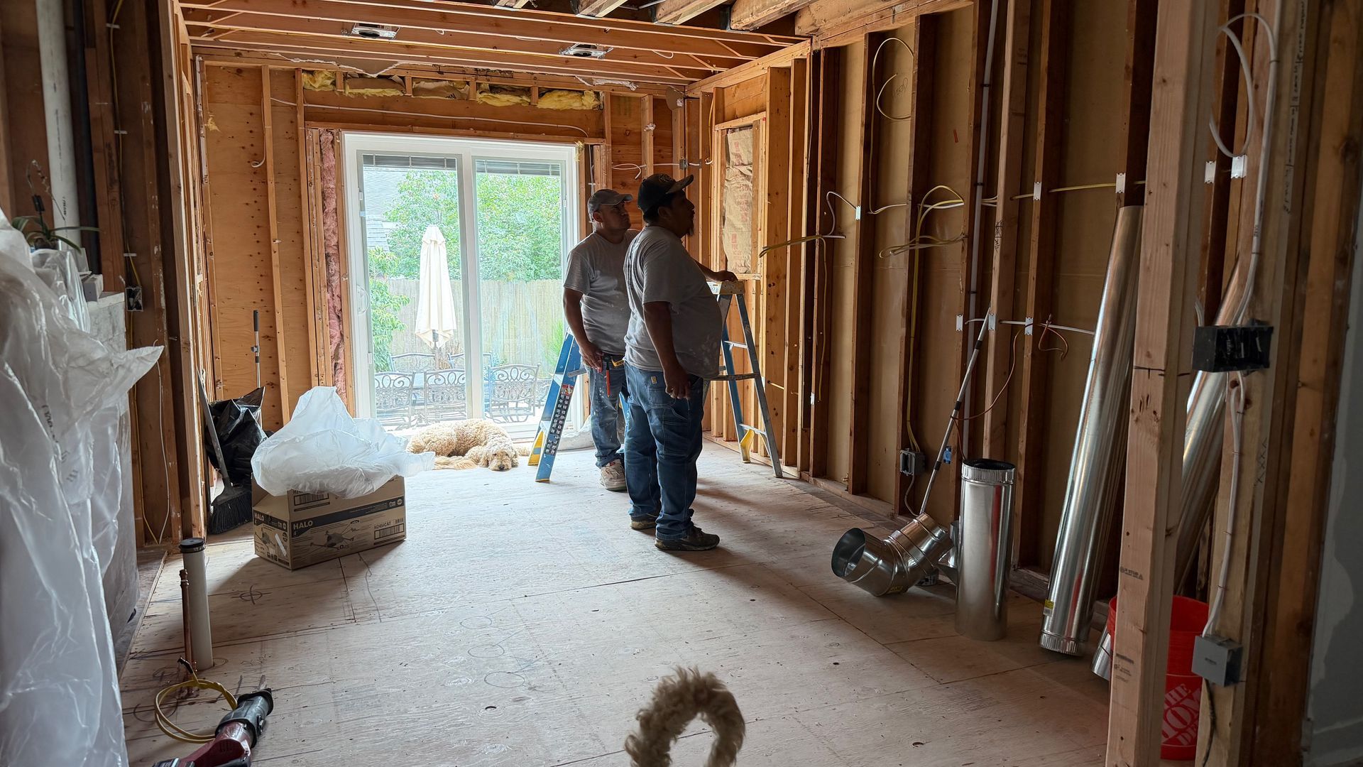 Two workers in an unfinished room with exposed studs and insulation, looking toward a bright doorway.