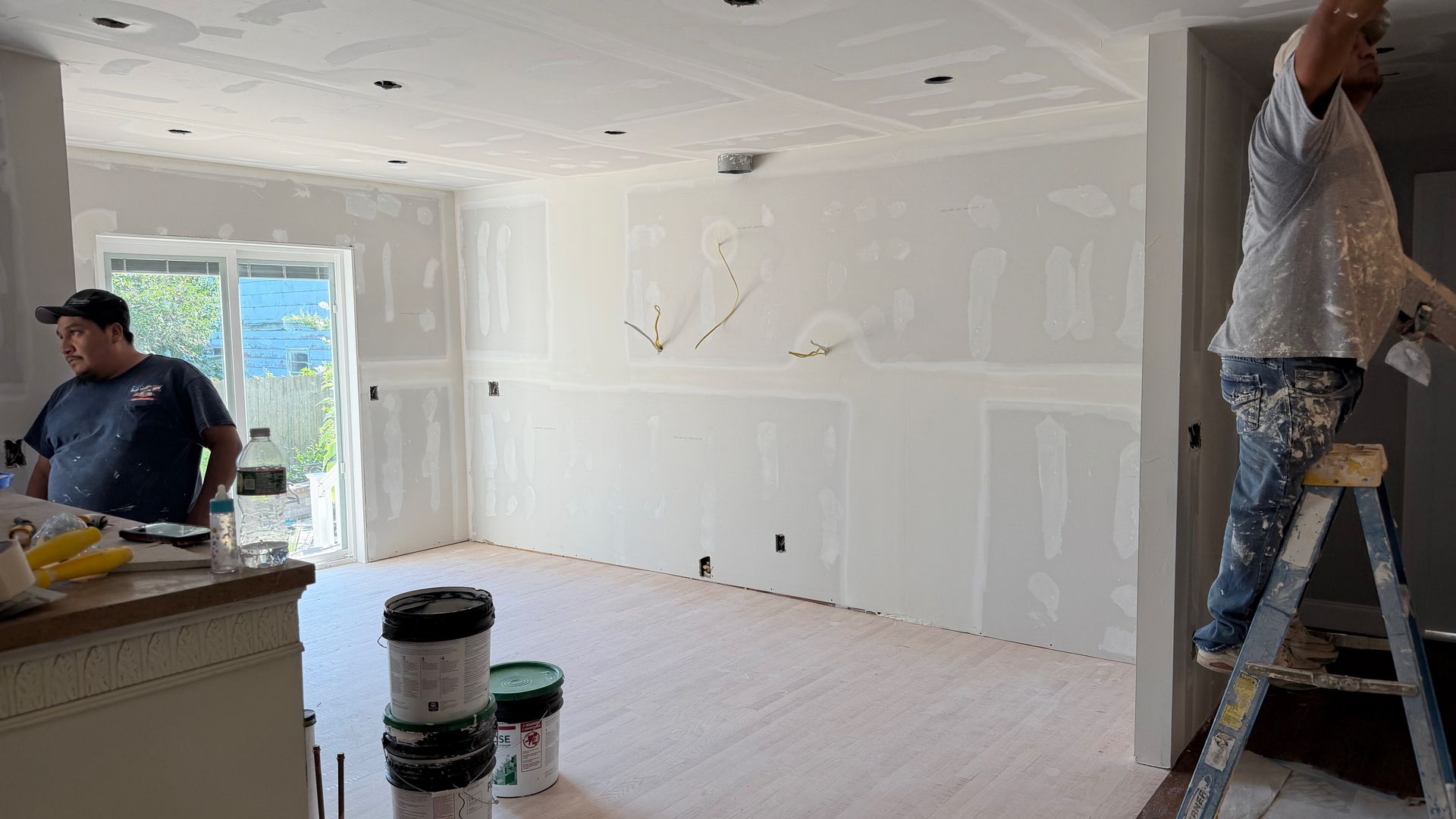 Two workers painting a bright white room under renovation, with ladders, tools, and covered floors.