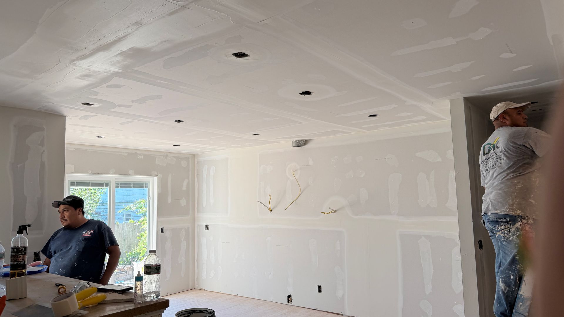 Partially finished room with drywall mudding, a man seated at left, and a worker in plastic at right.