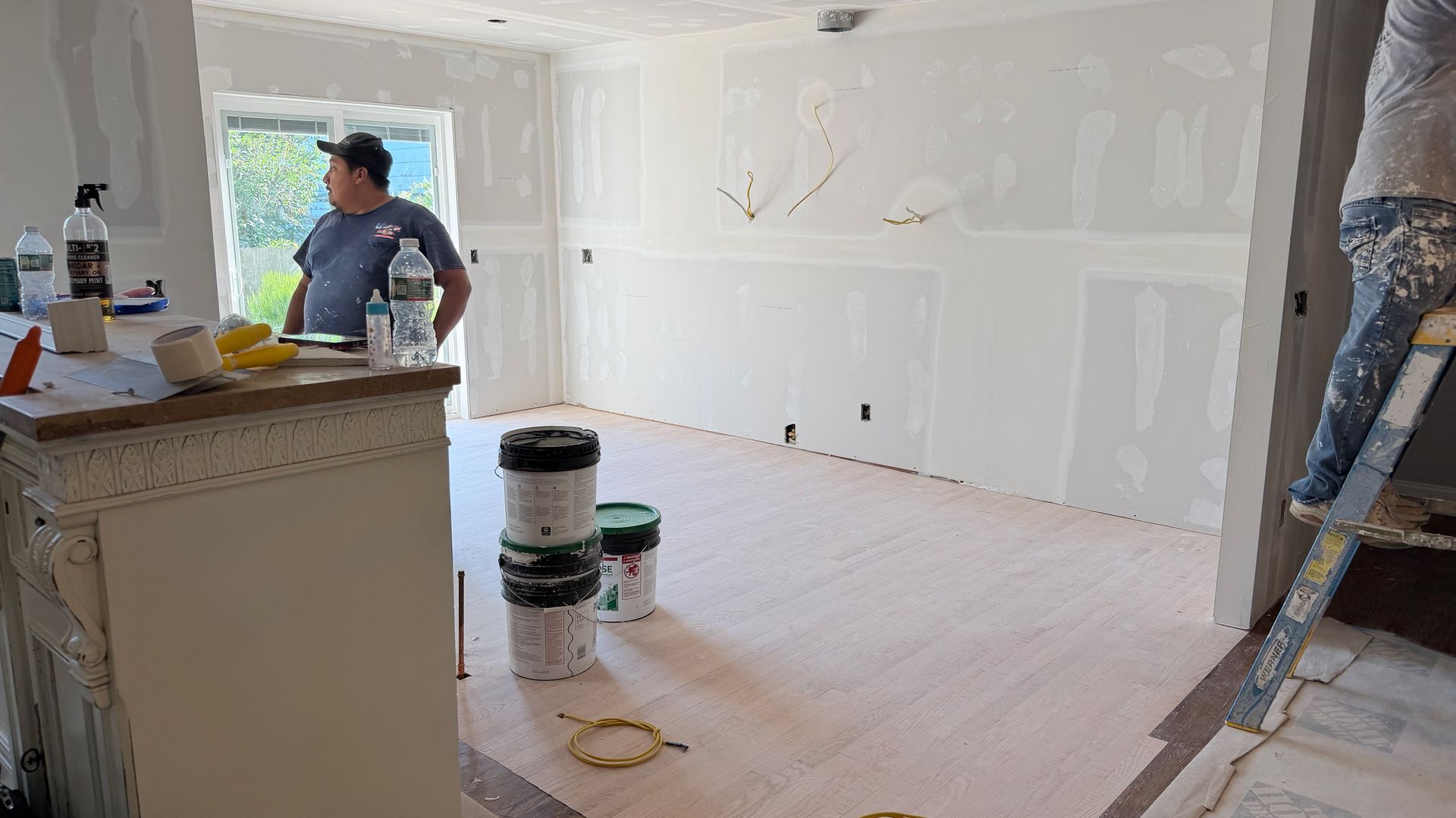 Partially finished room with white walls, a worker, paint buckets, and a ladder during renovation