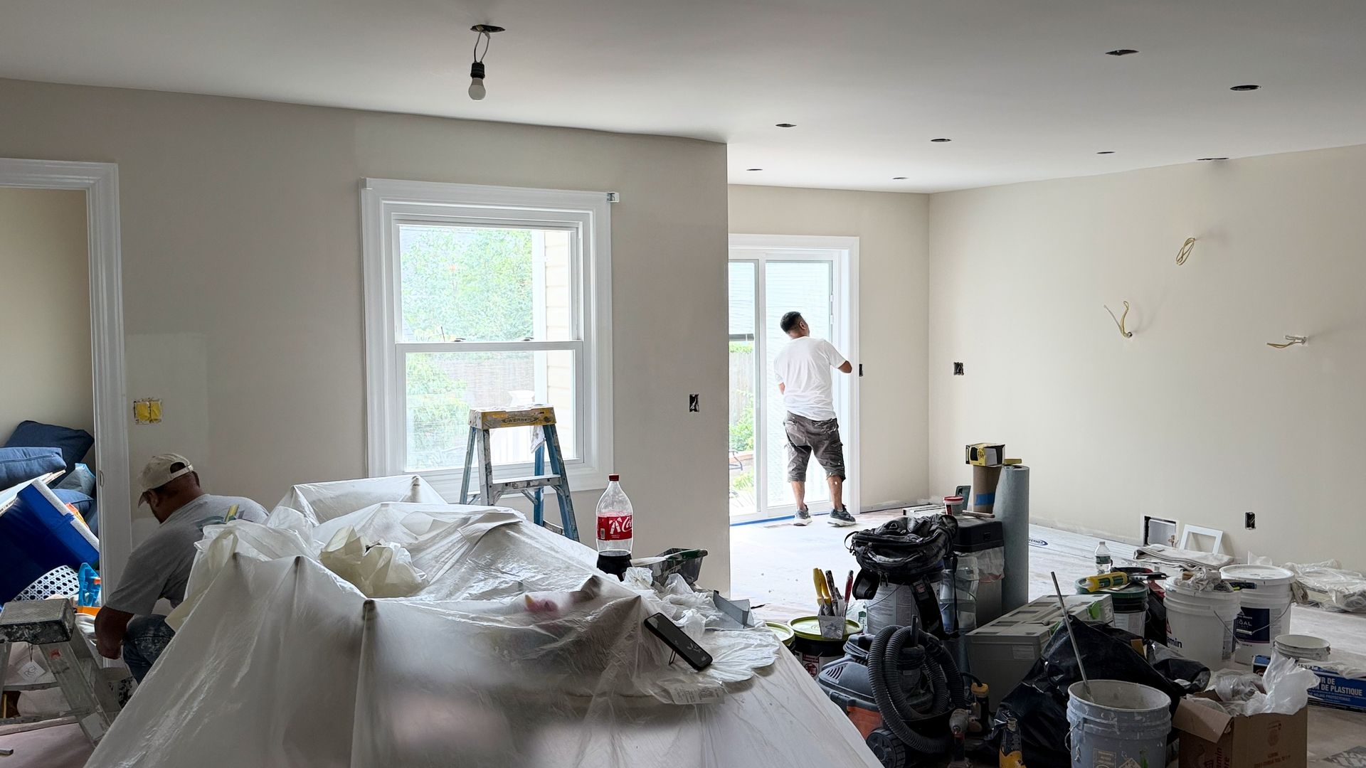 Bright living room under renovation with tools, debris, and a worker near the doorway.