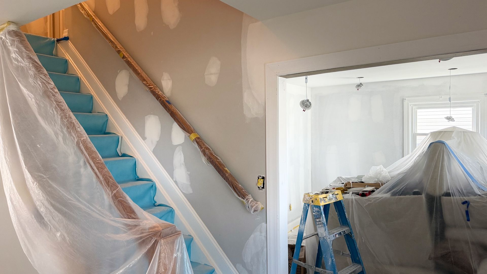 Staircase under renovation, covered in plastic, with blue carpet runner and a partially draped room beside it