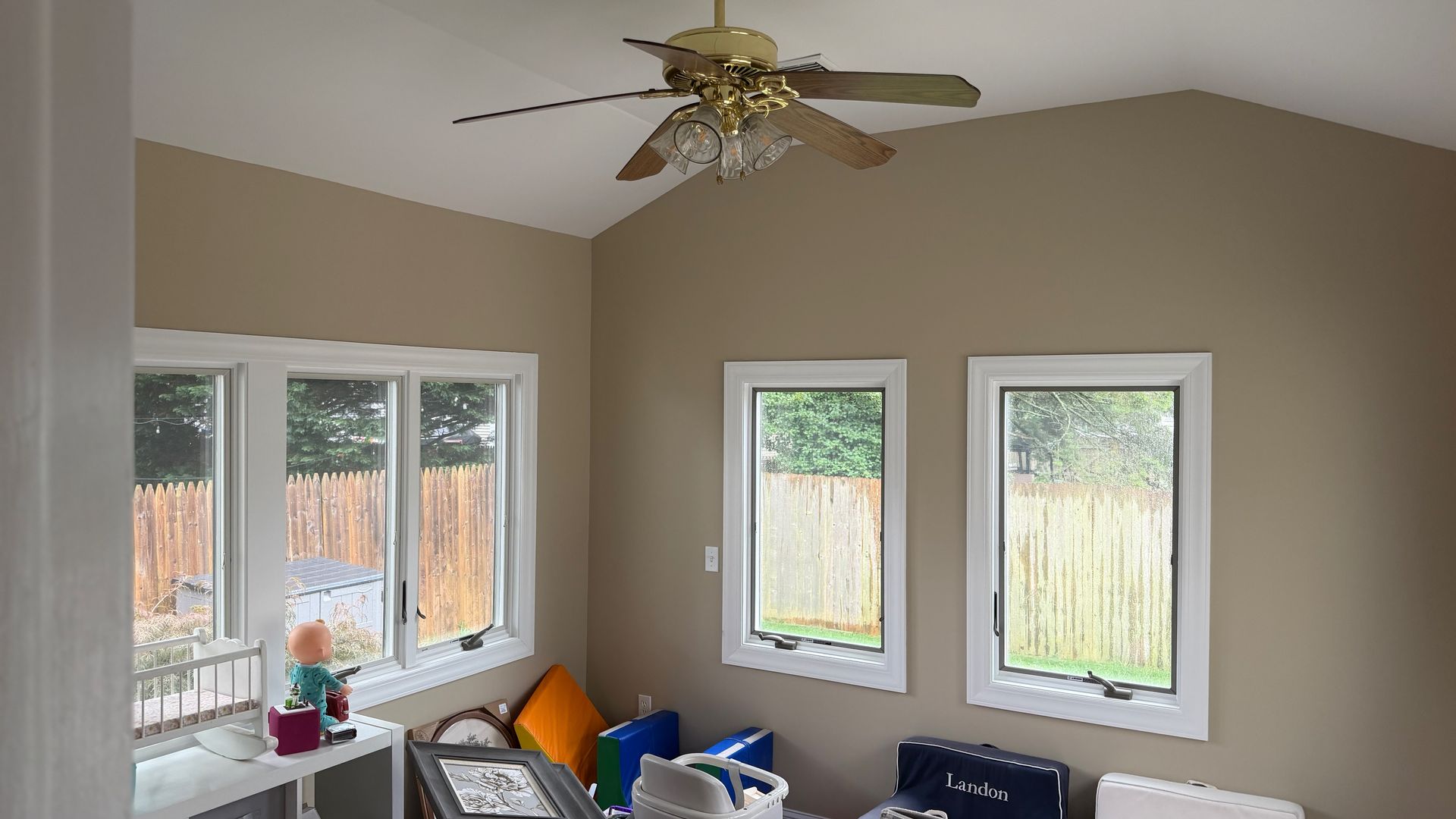 Small room with beige walls, white-trimmed windows, and a ceiling fan above a cluttered desk and chairs.