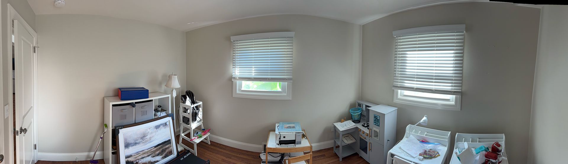 Bright nursery with a crib, changing table, two windows with blinds, and baby items on shelves.