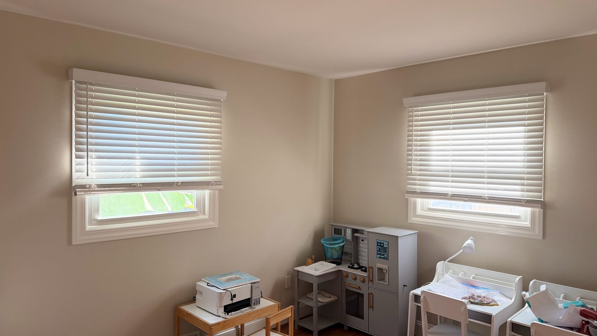 Small beige bedroom with two windows, blinds, and toys and storage furniture along the wall.
