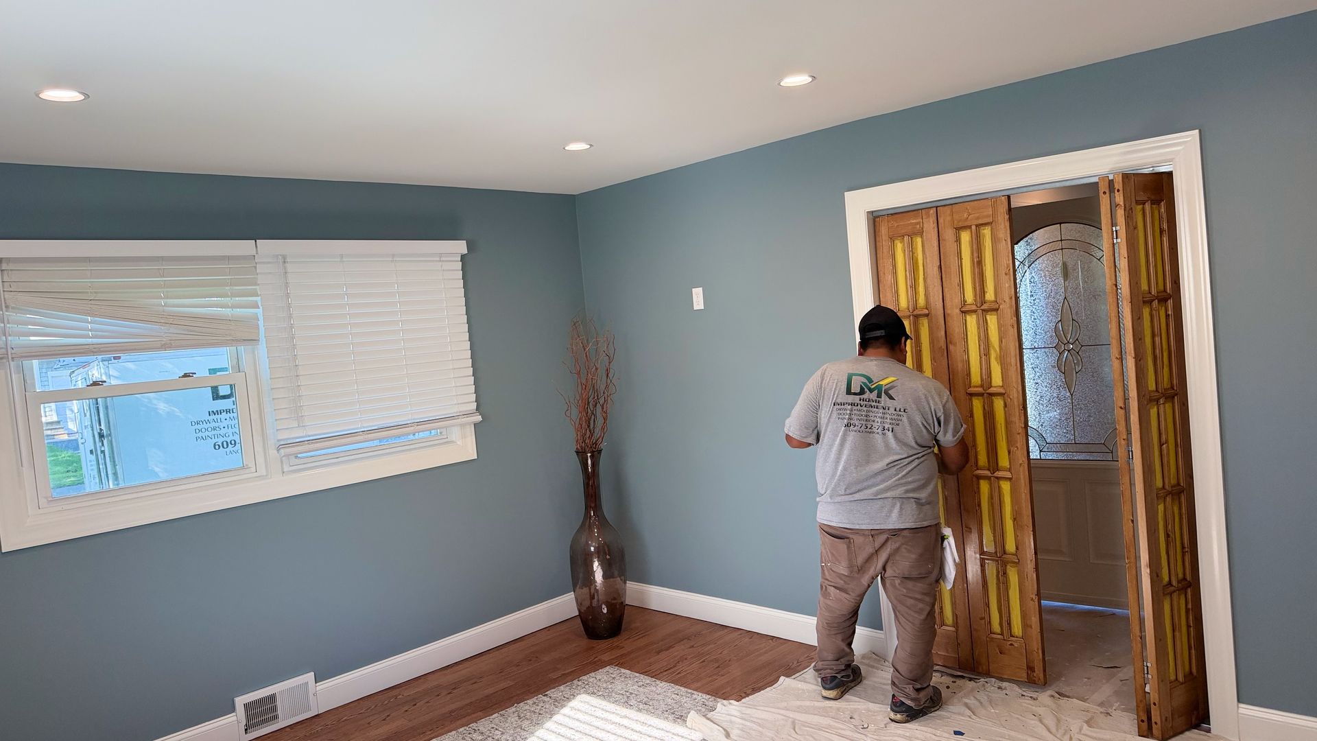 Person painting a wall in a blue room with white trim and a window.