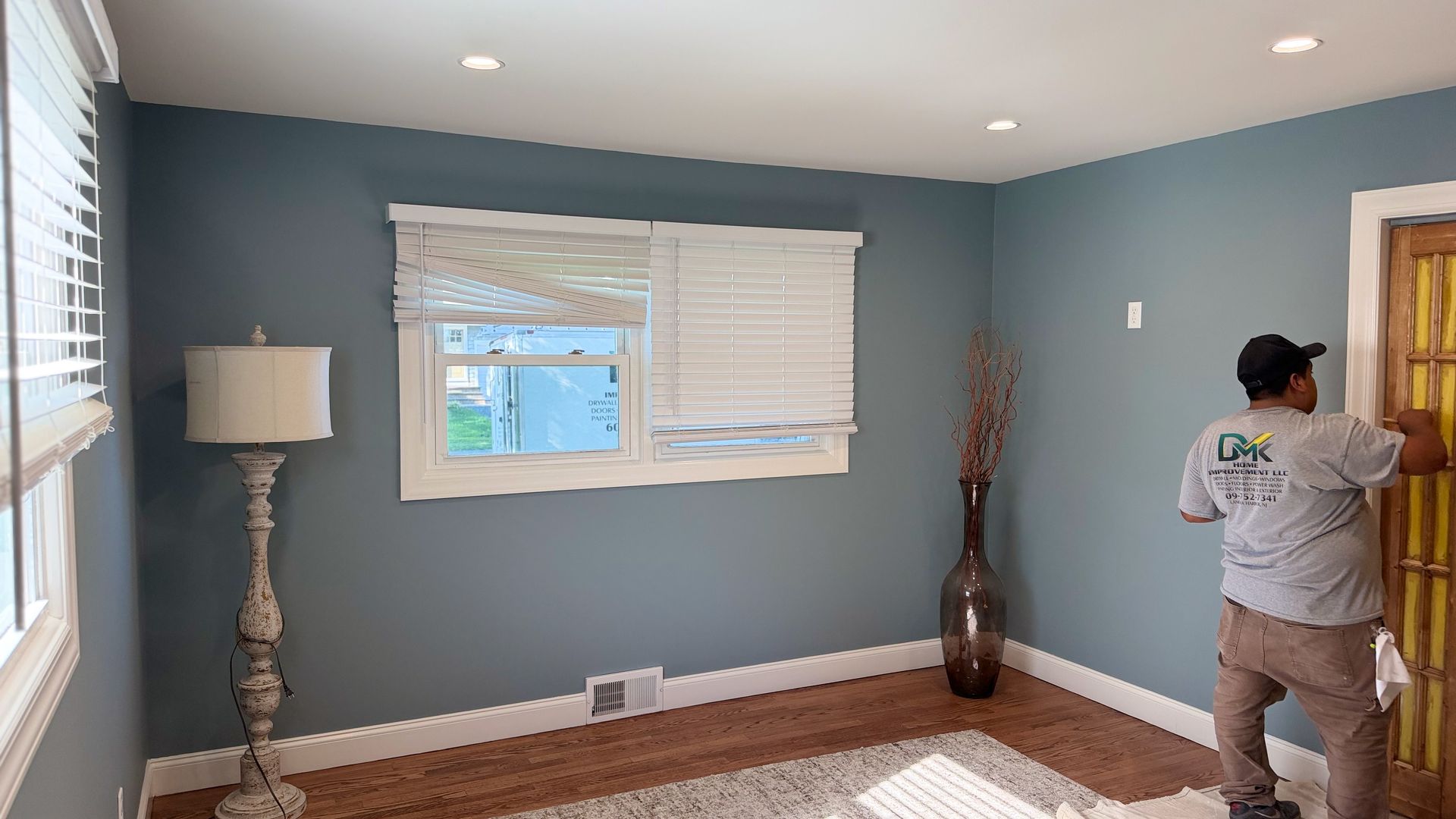 Empty blue room with window, floor lamp, and a person adjusting a wall on the right