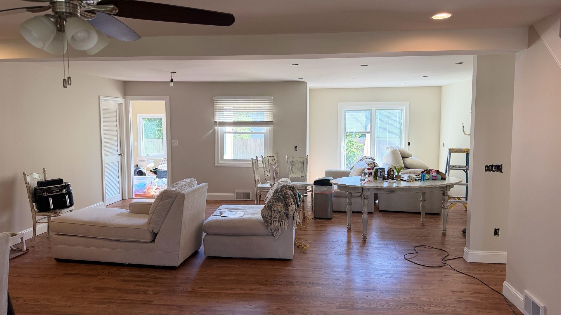 Bright living room with wood floors, white furniture, ceiling fan, and dining area in the background