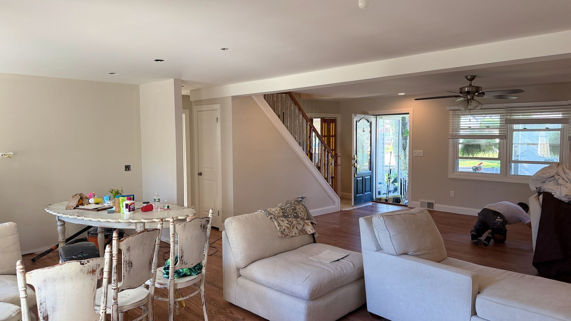Open-plan living room with white sofas, dining table, staircase, and a window letting in daylight.