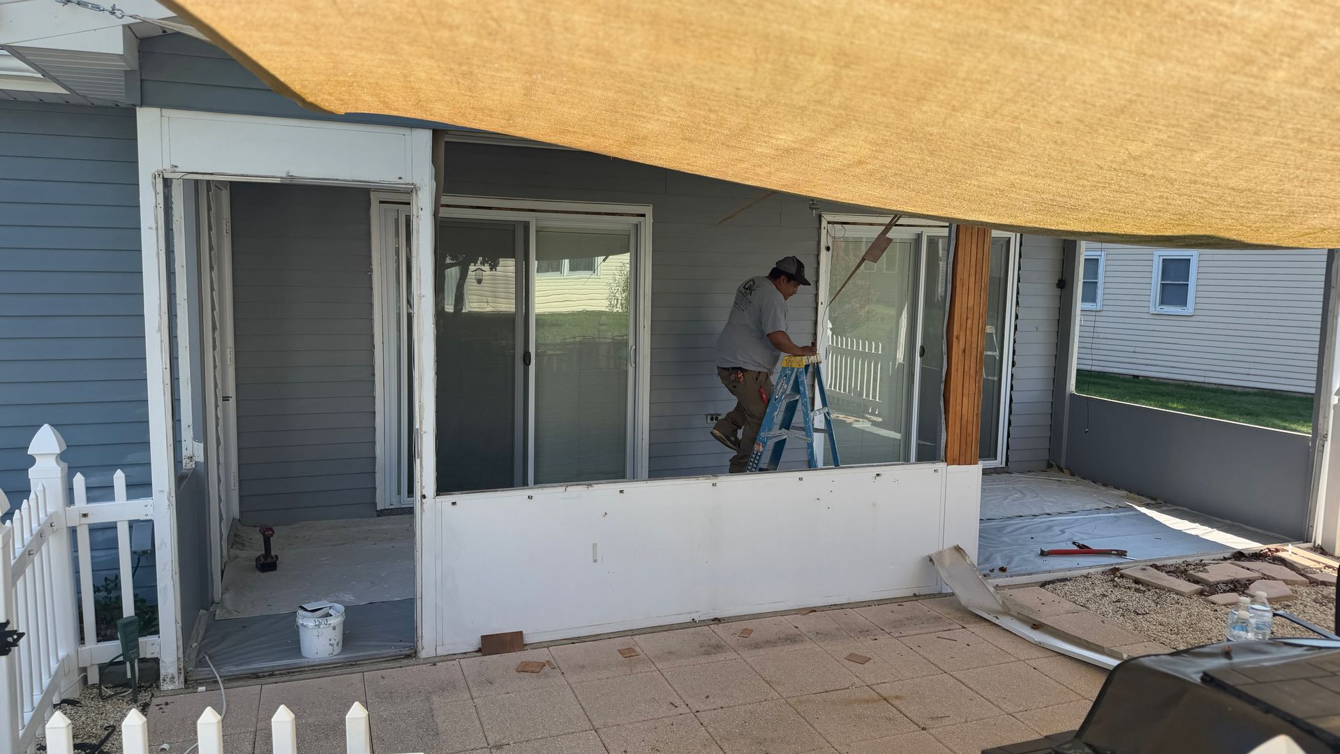 Worker repairing a covered porch with white siding and sliding glass doors under a tarp-covered roof