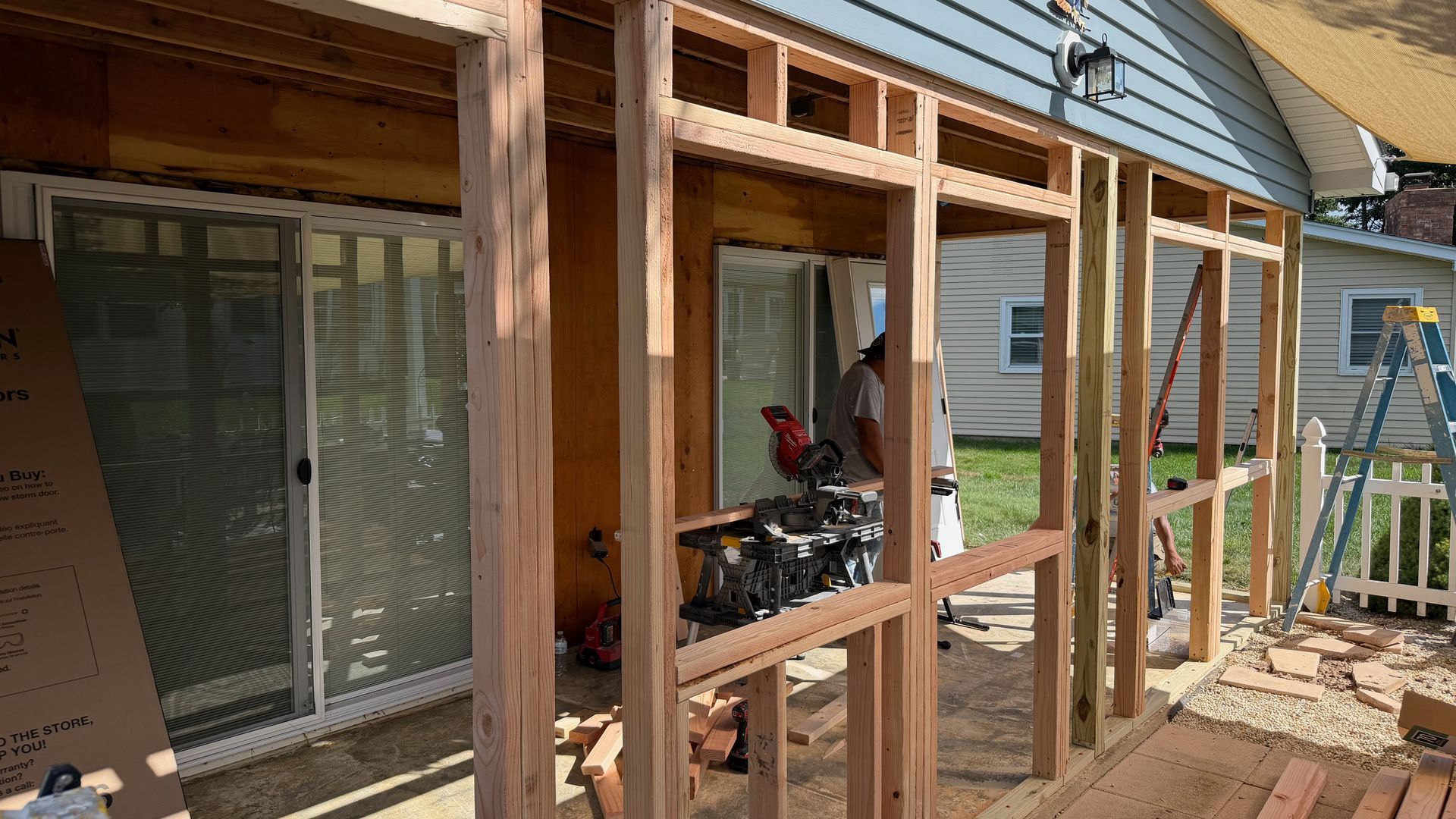 Wooden deck frame under construction beside a blue house, with tools and a ladder nearby.