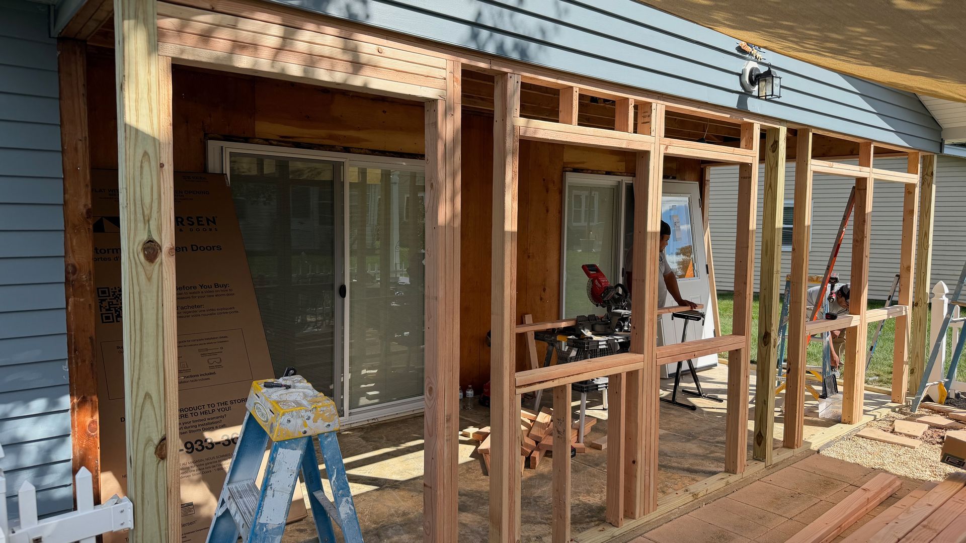 House exterior under renovation with exposed wooden framing, blue siding, and a ladder on the deck.